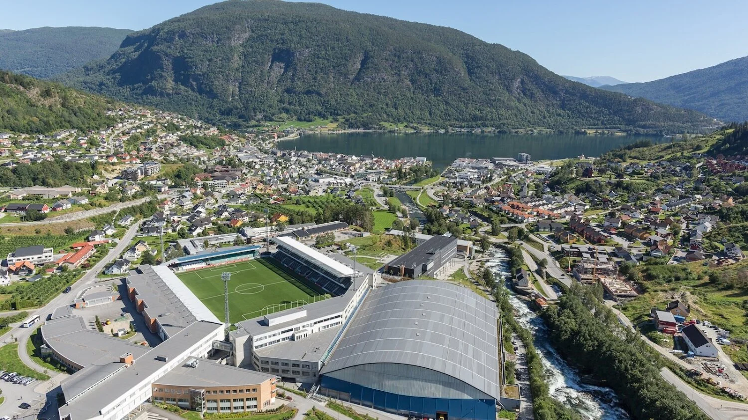 Bilde av en by med fjell og innsjø i bakgrunnen, og en fotballstadion foran.