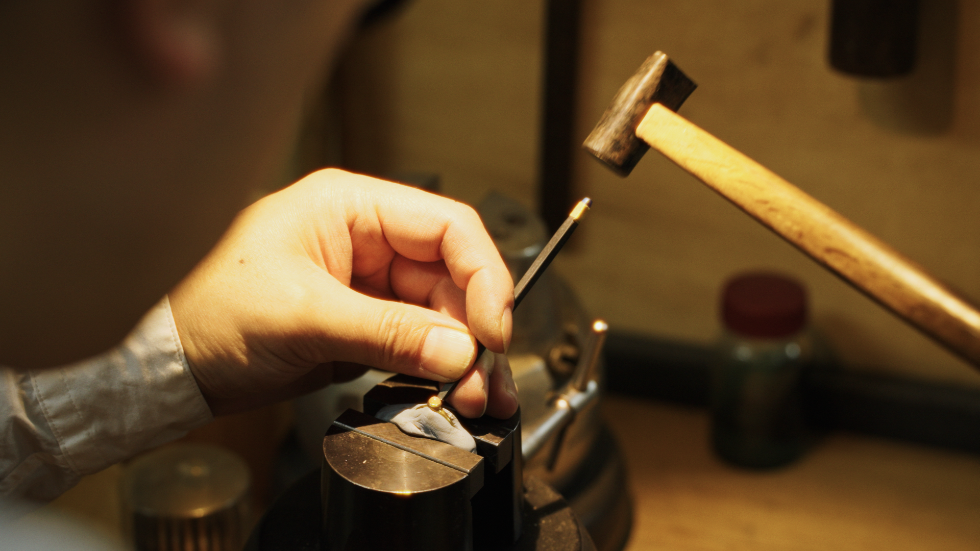 Close-up of a person's hand holding a small object with a tool in a workshop with a hammer and other tools in the background.