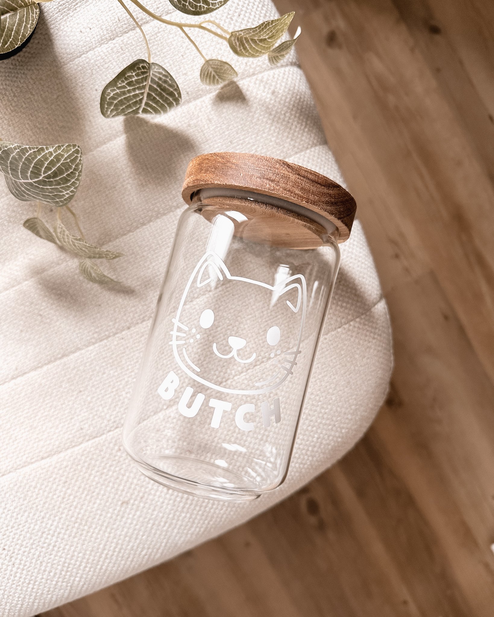 Un bocal en verre avec un couvercle en bois, décoré d'une illustration de chat et du mot 'BUTCH', posé sur une surface en tissu beige, avec une plante à feuilles vertes en arrière-plan.