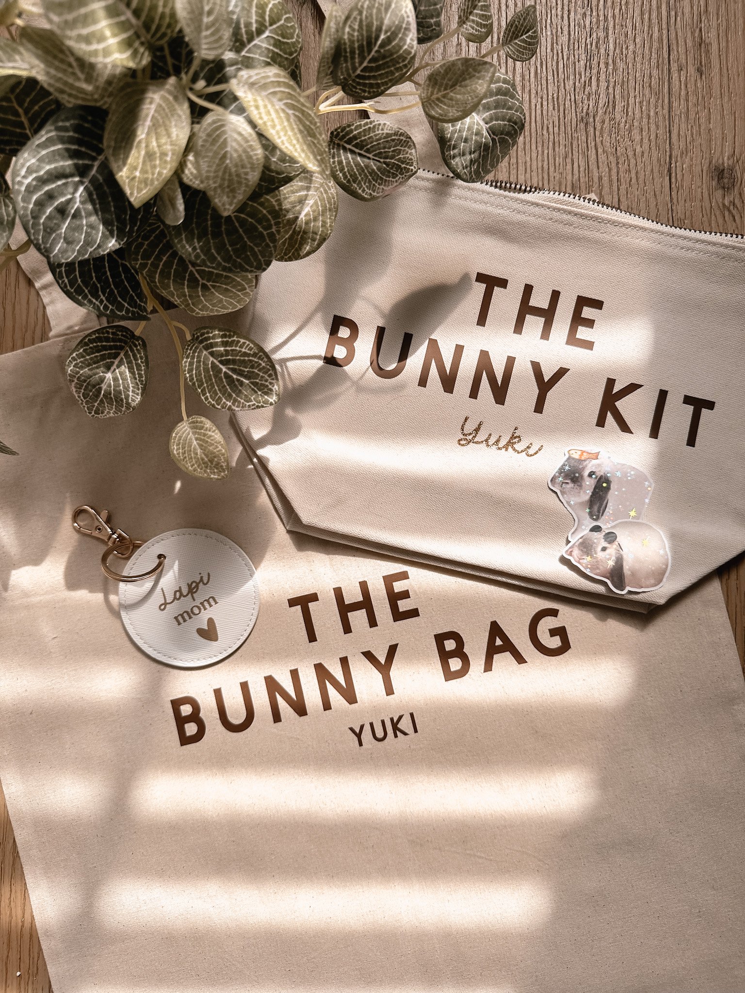 Une pochette blanche avec le texte 'The Bunny Kit Yuki'. Un sac en tissu beige avec le texte 'The Bunny Bag Yuki'. Une plante verte en haut à gauche. Une clé en forme de cercle avec 'Lapi mom' et un cœur, posé sur la pochette.