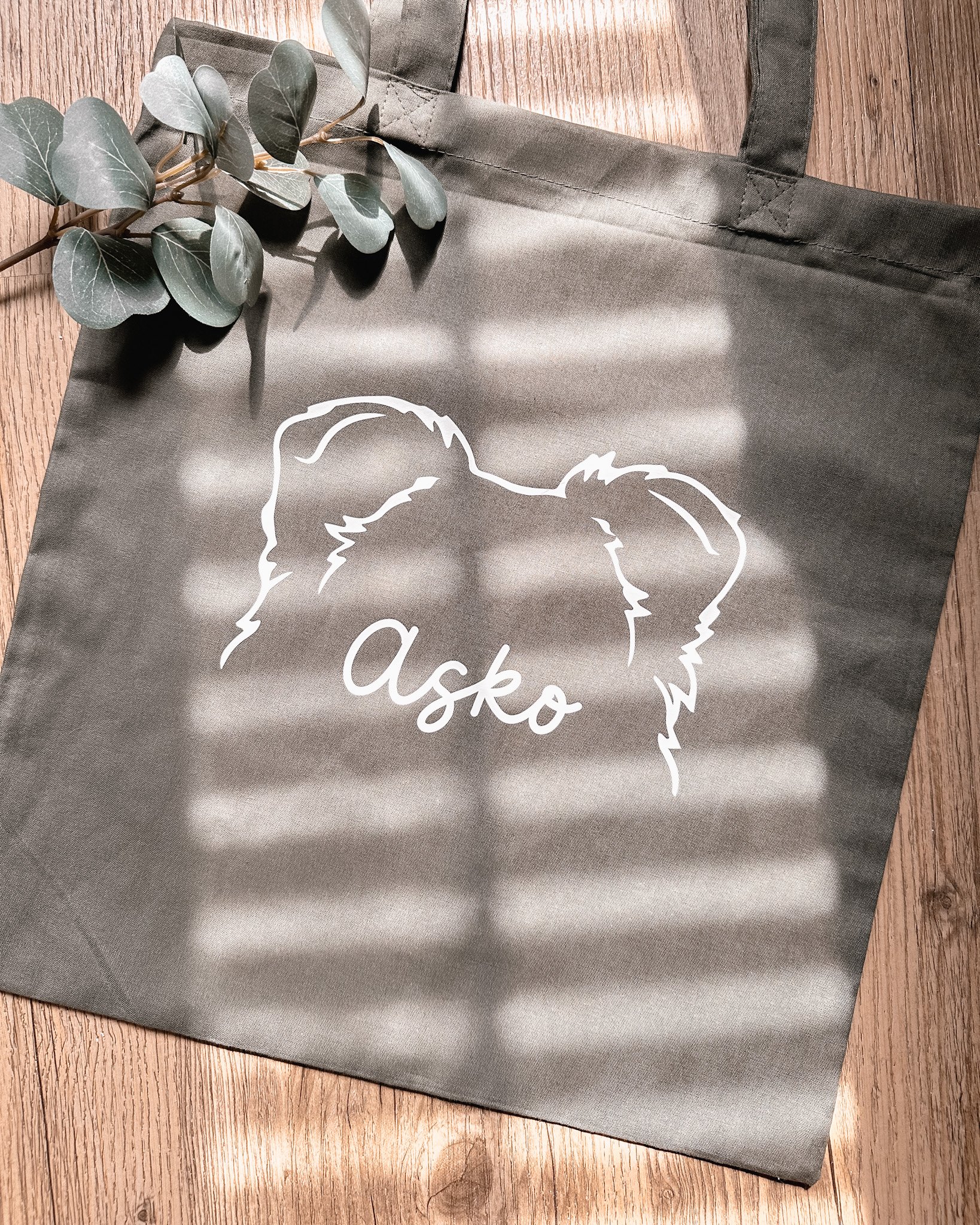 Un sac en tissu gris clair avec une illustration de tête d'ours en ligne blanche et le nom 'Asko' écrit en cursive. Sur le sac, il y a une branche de feuilles vertes et des rayures de lumière qui tombent dessus, reposant sur un sol en bois.