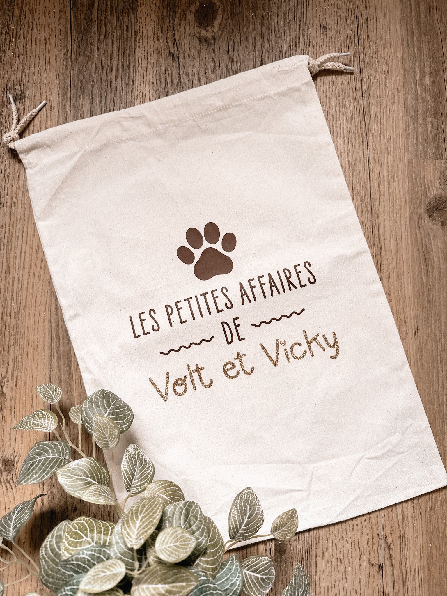 Sac en tissu avec écritures en français et une empreinte de patte de chien, placé sur un sol en bois, avec des feuilles vertes en premier plan.