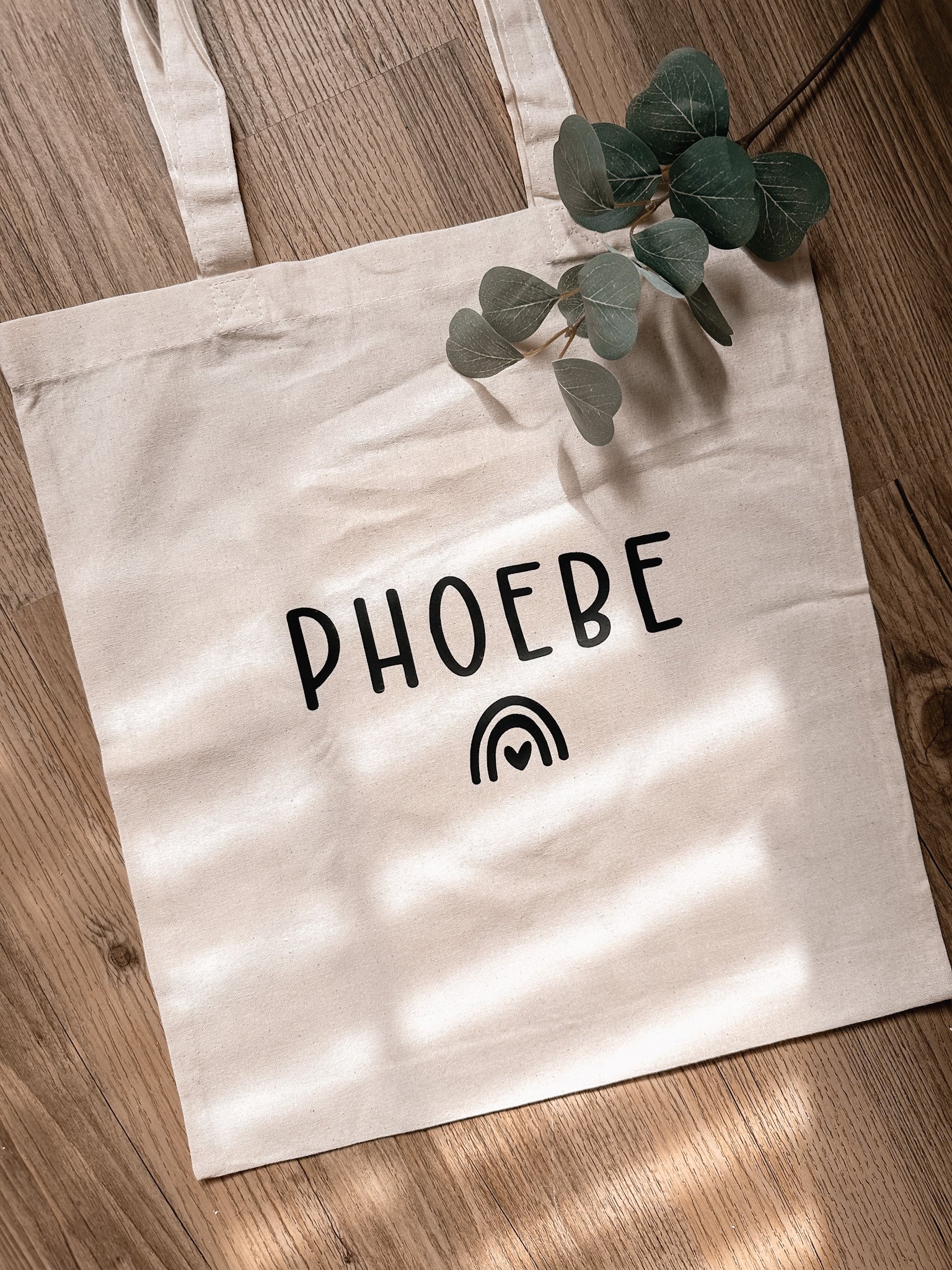 Sac en toile beige avec le nom 'PHOEBE' écrit en noir, décoré d'une petite illustration d'un arc-en-ciel avec un cœur, posé sur un plancher en bois avec une branche de feuilles vertes à côté.