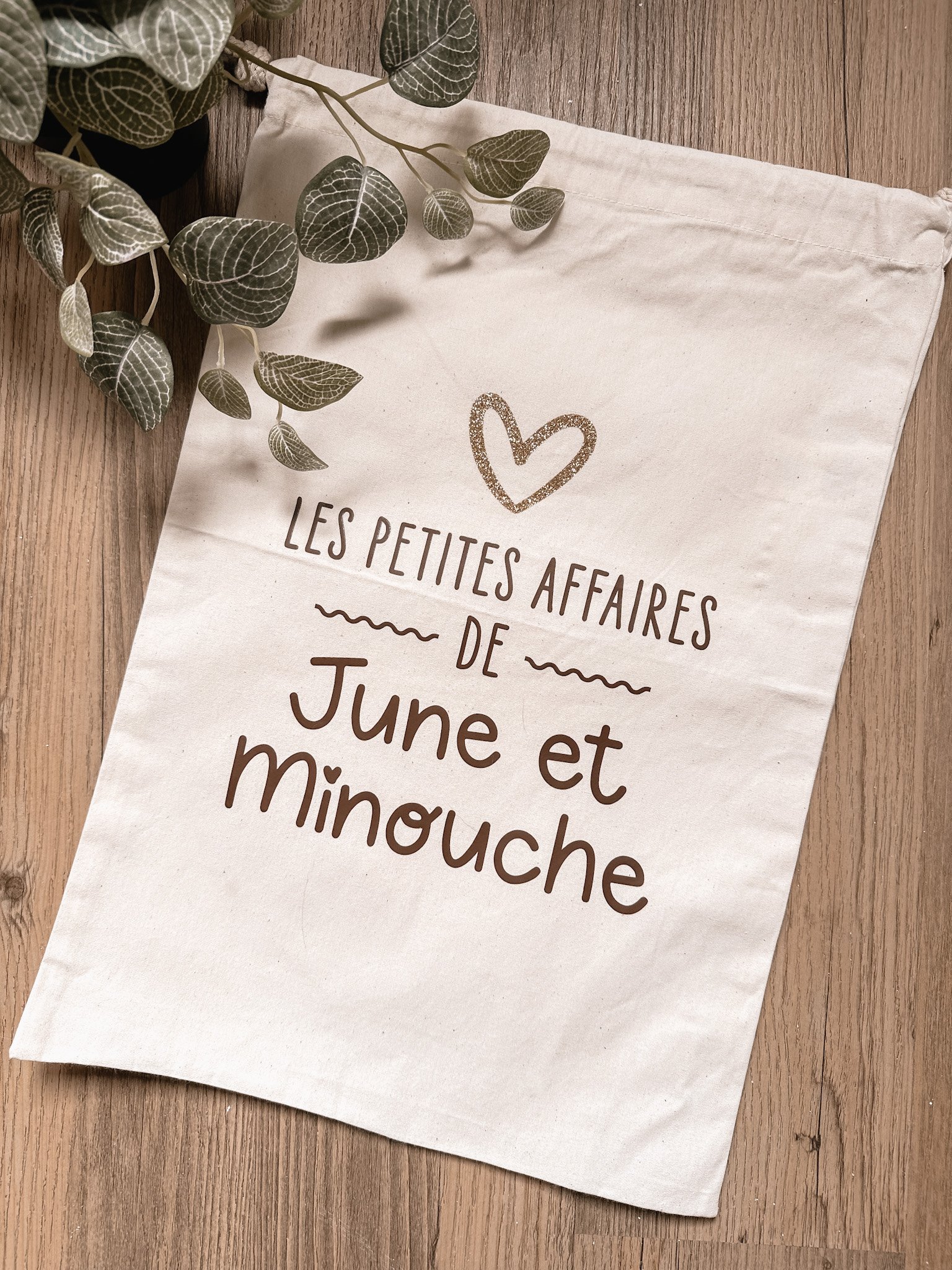 Une bannière en tissu avec le texte "Les petites affaires de June et minouche" accompagnée d'un dessin de cœur, posée sur un plancher en bois avec des feuilles vertes en haut à gauche.