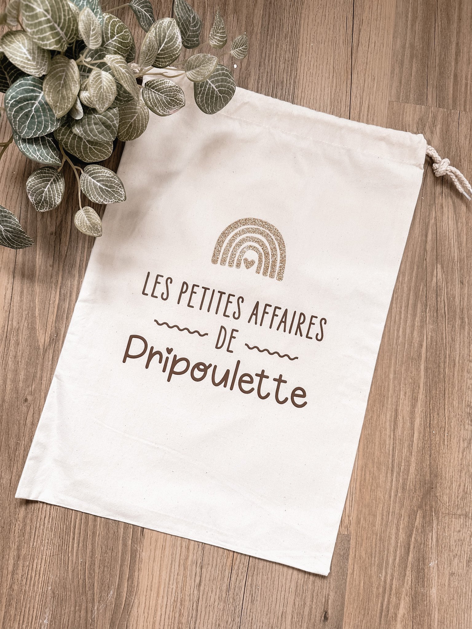 Sac en coton blanc avec inscription en français et dessin d'arc-en-ciel, posé sur un sol en bois, à côté d'une plante verte.