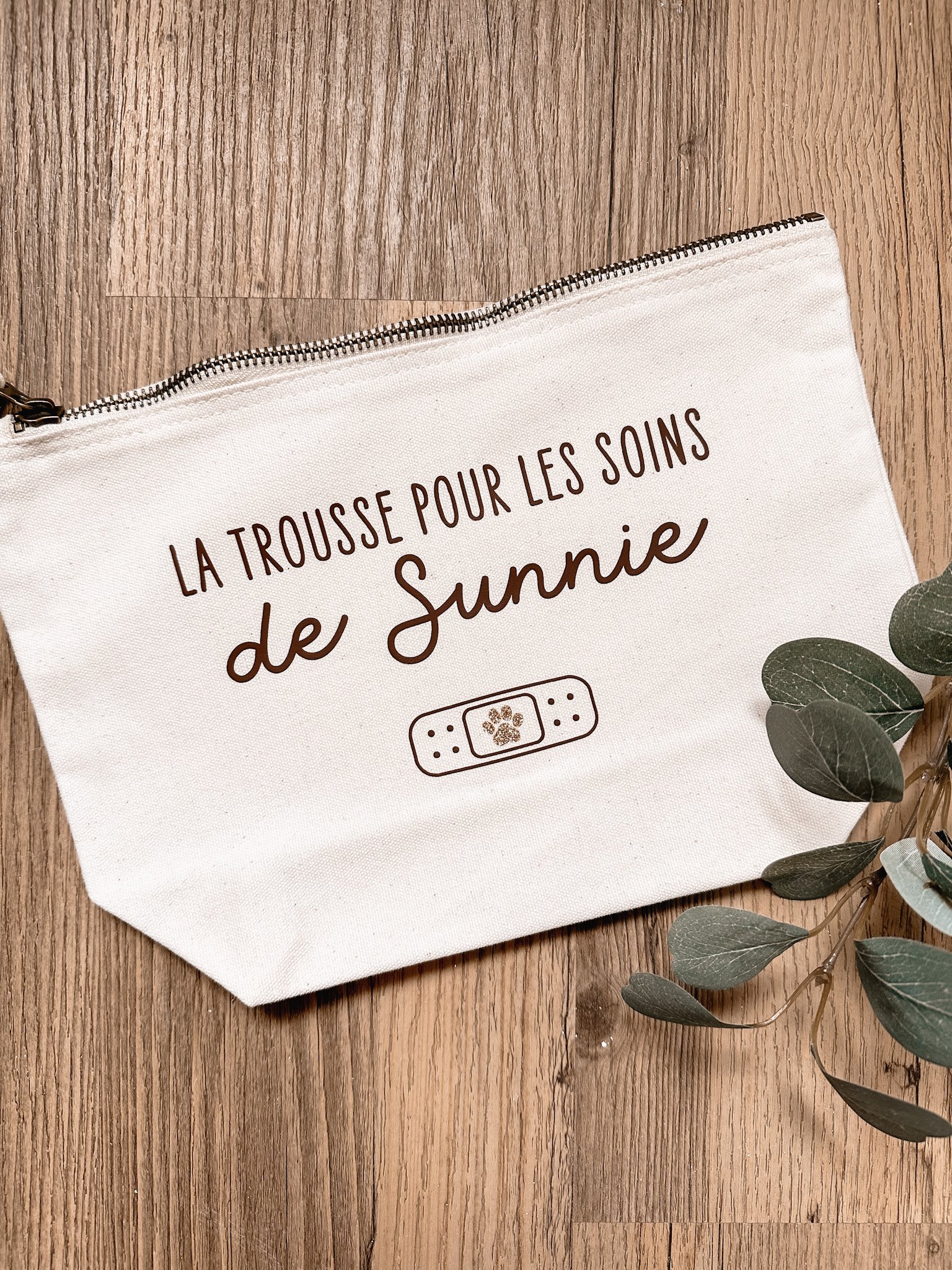 Une trousse blanche avec le texte 'La trousse pour les soins de Sunnie' posée sur un sol en bois, avec quelques feuilles vertes à droite.