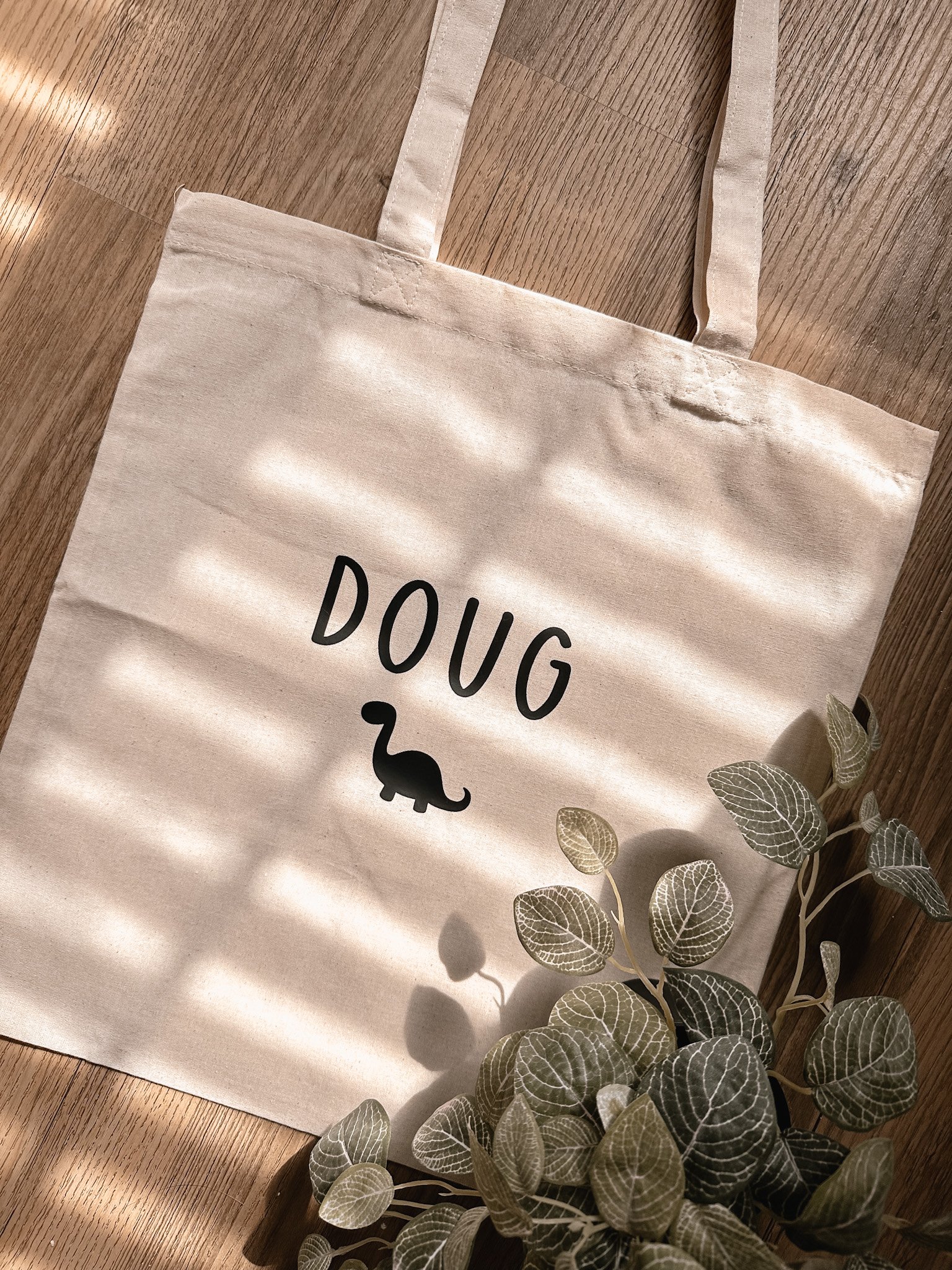 Un sac en toile beige avec le mot 'DOUG' écrit dessus et une icône de dinosaure noir, posé sur un plancher en bois, avec une plante verte en première plan.