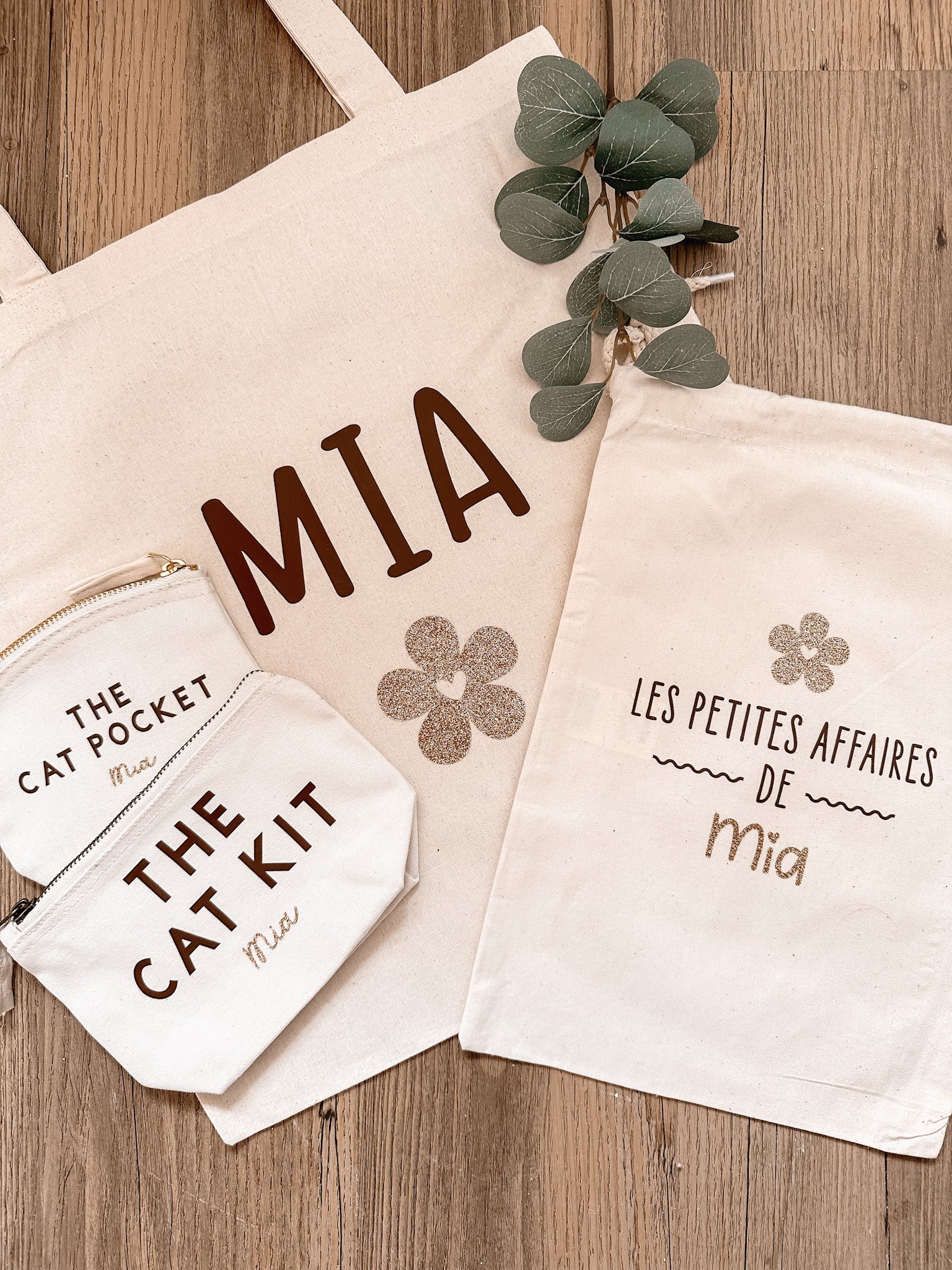 Tissus en coton beige avec écritures pour Mia, comprenant une pochette blanche avec l'inscription 'The Cat Pocket', un sac en tissu avec 'Les Petites Affaires de Mia' et un sac avec 'The Cat Kit', décorés de fleurs dorées et de branches de eucalyptus