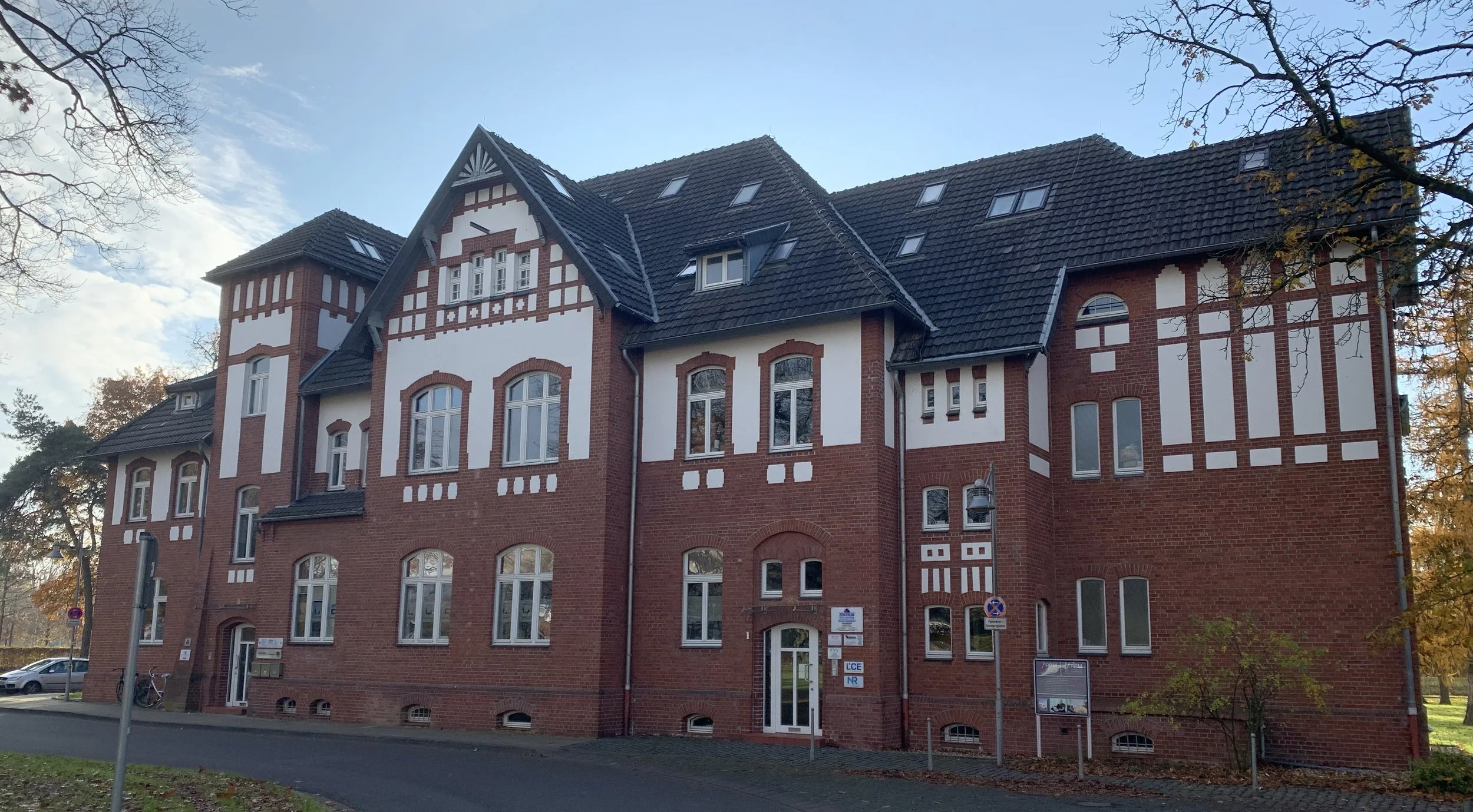 Historisches rotes Backsteingebäude mit weißen Putzdetails, mehreren Fenstern, Dächern mit Gauben, im Hintergrund blauer Himmel und Laubbäume im Herbst.