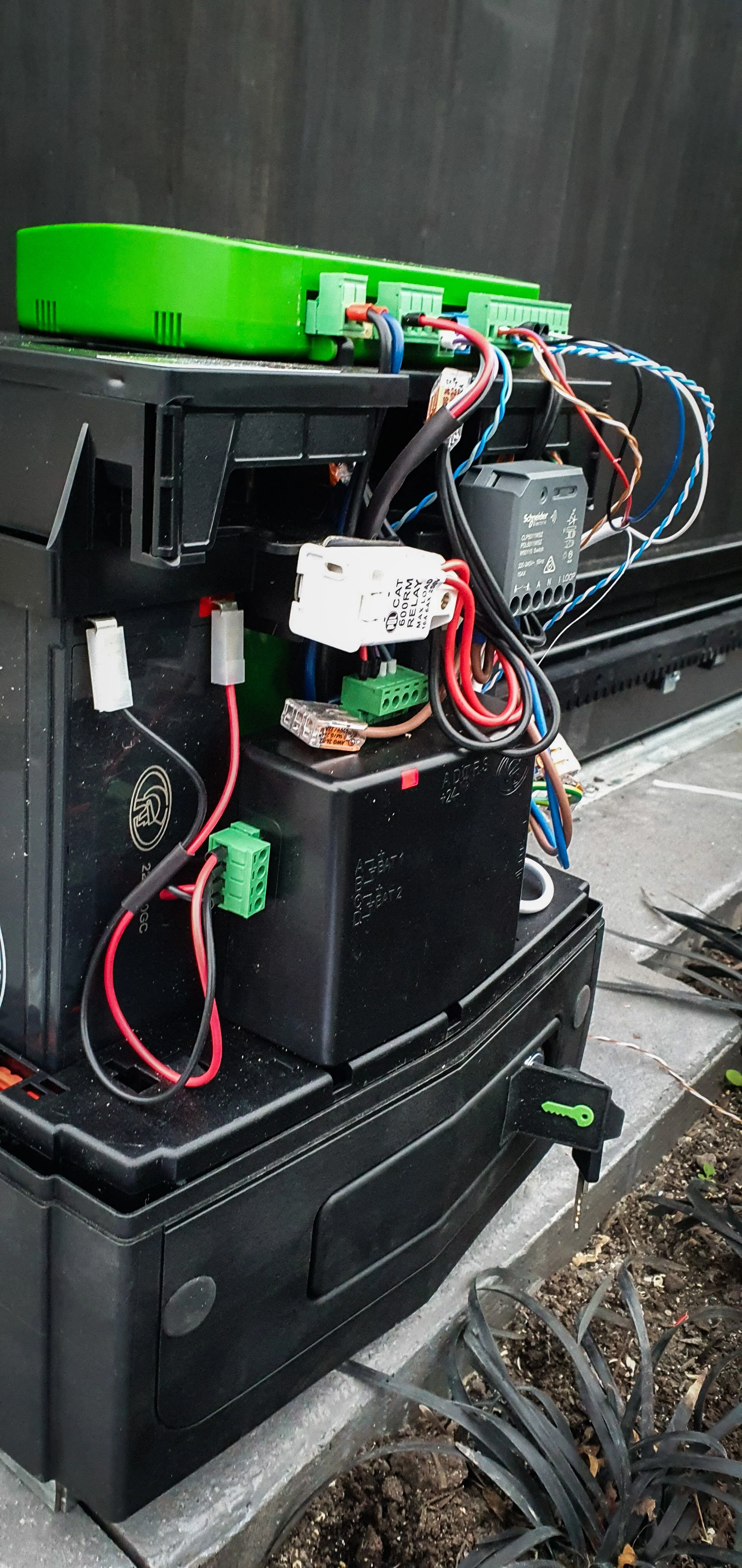Close-up of an outdoor electrical control box with wires, relays, and circuit boards, mounted on a black plastic enclosure, with soil and plants nearby.