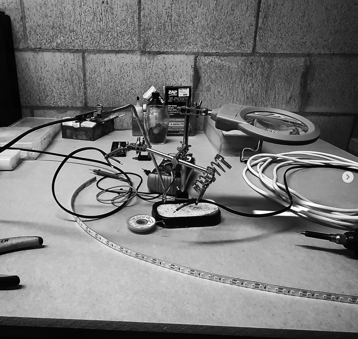 A cluttered workbench with soldering equipment, wires, tools, and small containers against a cinder block wall.