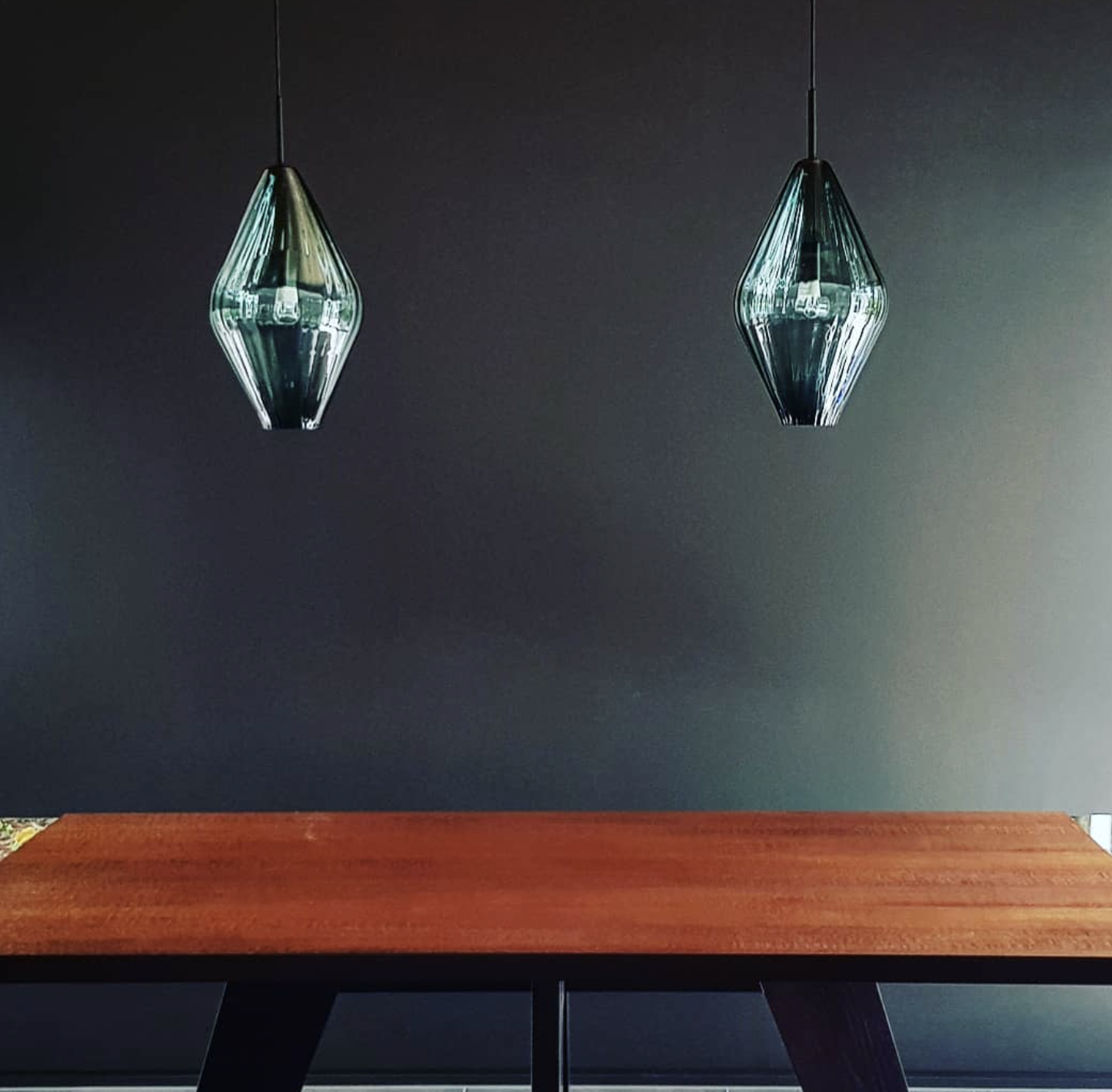 Two teal glass pendant lights hanging above a wooden table against a dark wall.