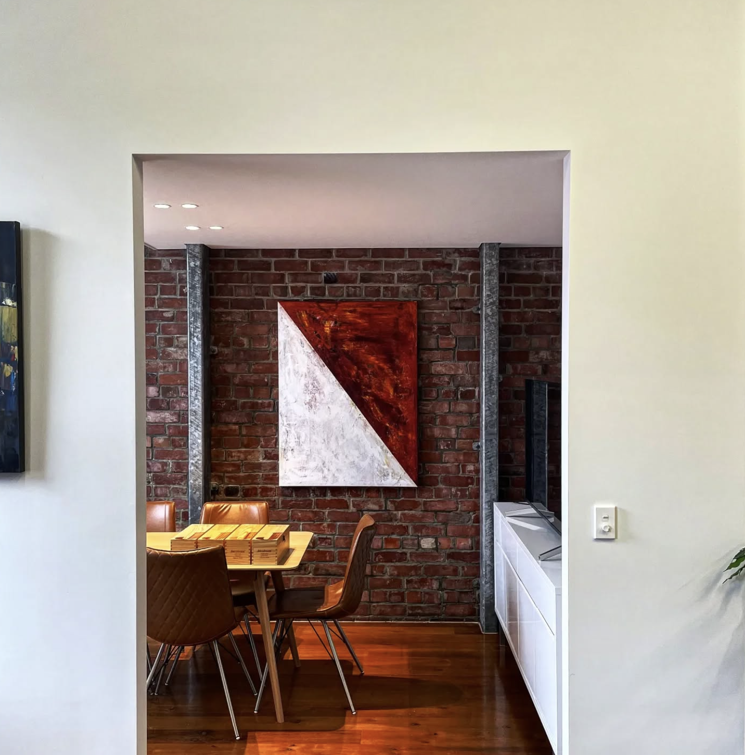 View of a dining area with a wooden table and brown chairs, a brick wall with an abstract art piece, and a white cabinet with a television on it.