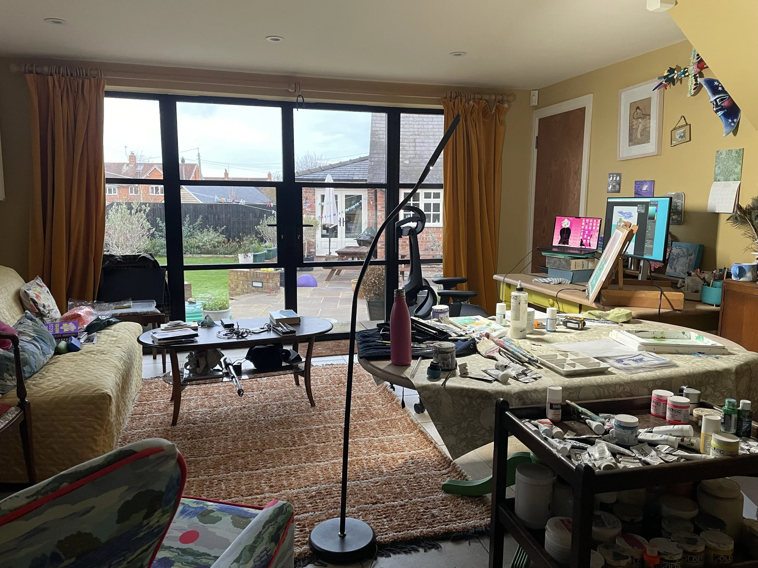 A living room with a large window overlooking a backyard, cluttered with art supplies, a computer, a desk, a sofa, a small table, and a rug.