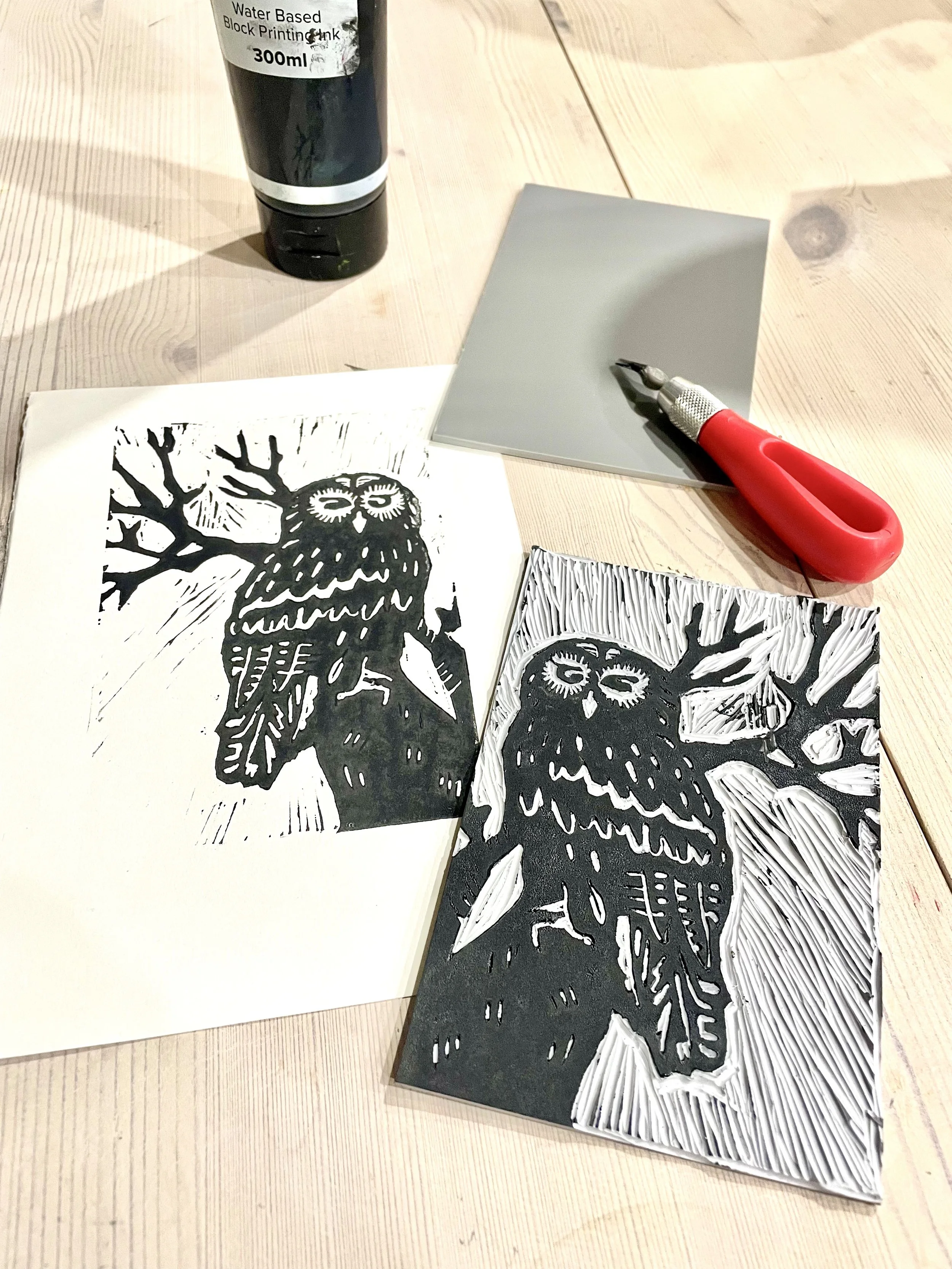 Black and white linocut print of an owl on a tree branch, with carving tools and a water-based block printing ink bottle on a wooden table.