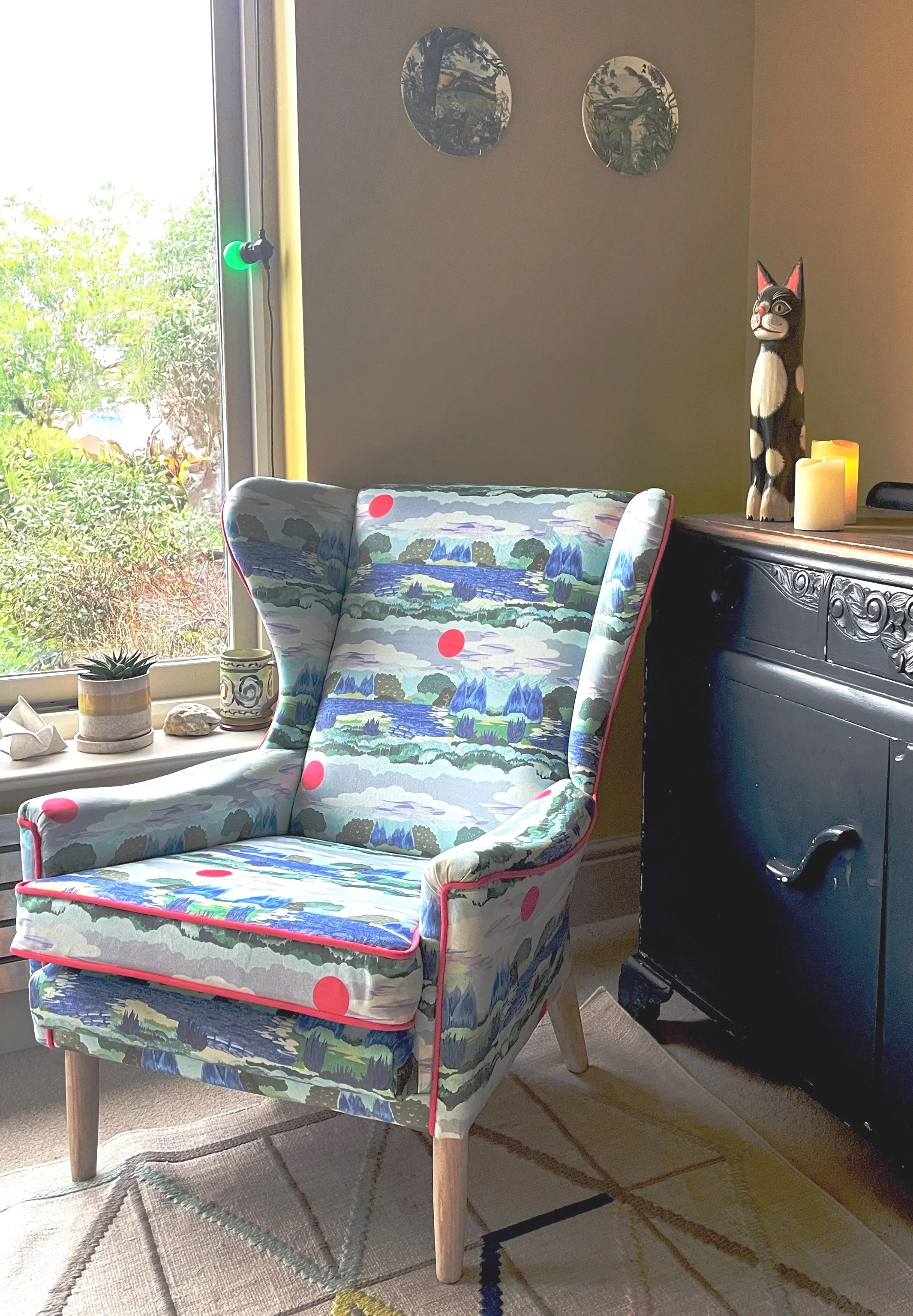 A living room corner with a colorful patterned armchair, a black sideboard with a cat-shaped sculpture, candles, and small plants on the window sill. A window shows greenery outside.