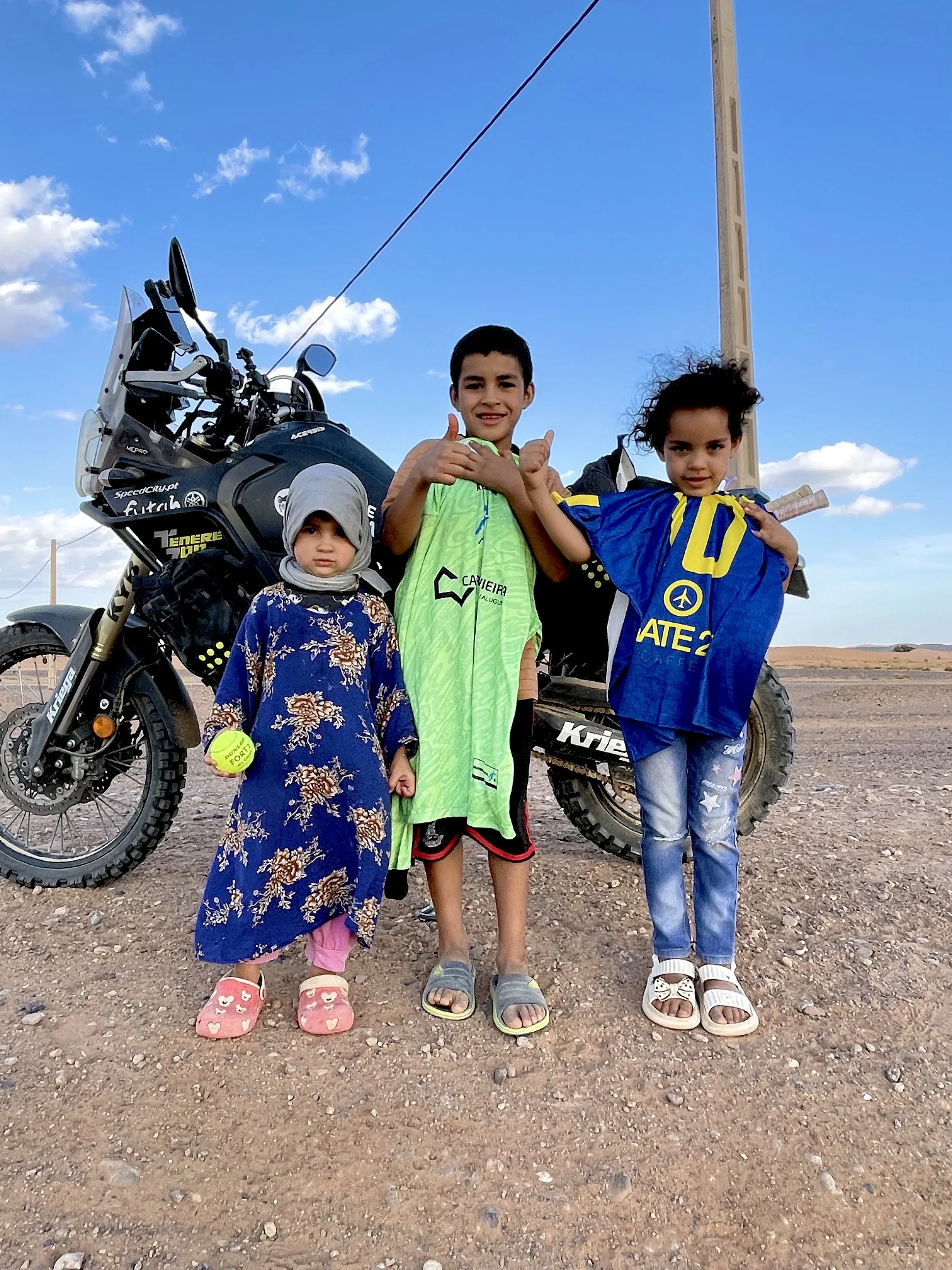 Yamaha Ténére 700 motorcycle with a group of little kids holding a tennis ball, and sport clothes donated by Decathlon, in Merzouga during a Beyond T7nnis Mission