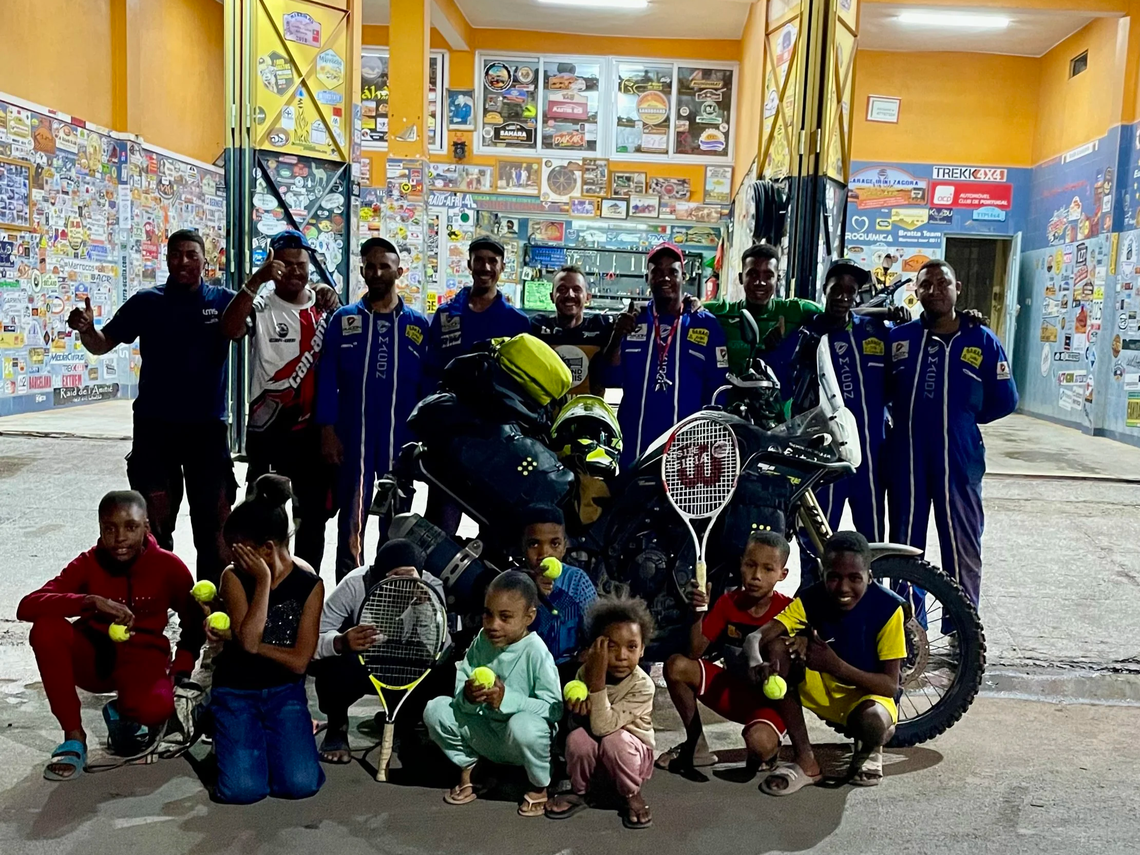 João Beltrão founder of Beyond T7nnis with his Yamaha Ténére 700 motorcycle with a group of kids and mechanics from Iriki Garage in Zagora, Morocco,  holding donnated tennis rackets and balls during a Beyond T7nnis Mission