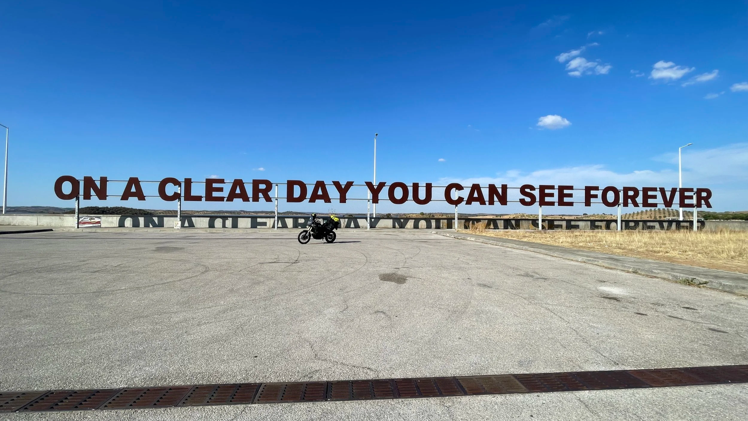 A Yamaha Ténéré 700 motorcycle with Kriega luggage on Alqueva Dam in Portugal. Big sign board with On a clear day you can see forever