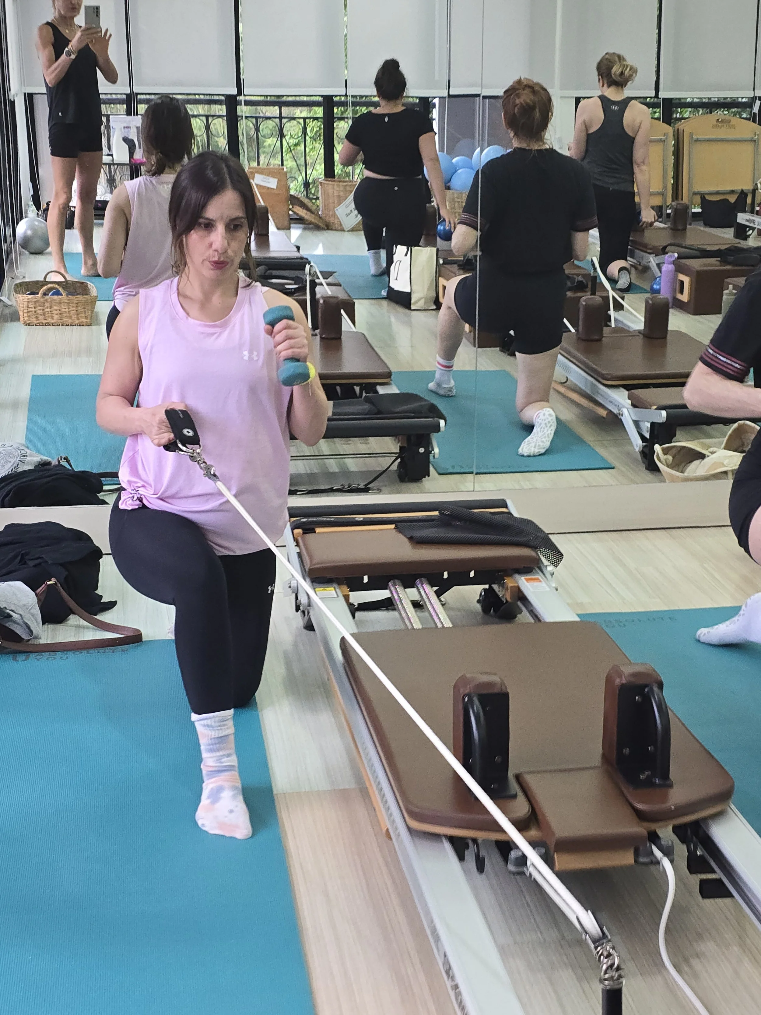 A woman in a pink tank top and black leggings stretching her leg on a Pilates reformer machine in a fitness studio with mirrored walls and other women exercising in the background.