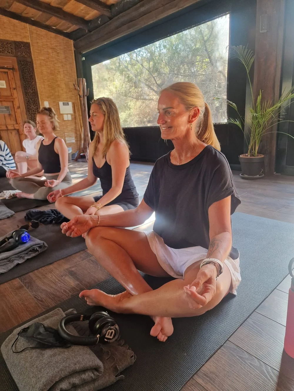 A group of women sitting cross-legged on yoga mats in a peaceful indoor setting, practicing meditation, with a woman in the foreground smiling.