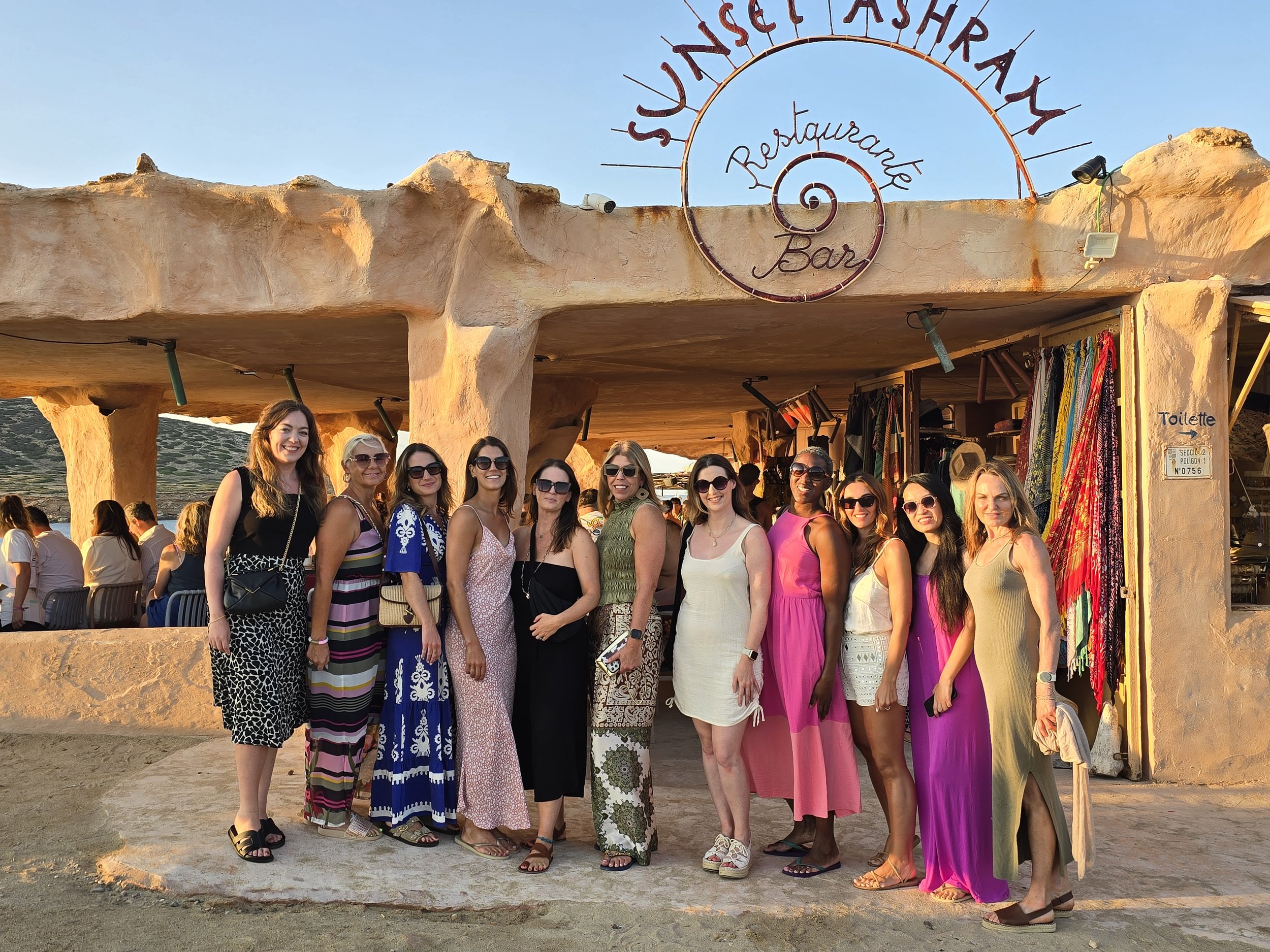 A group of women standing in front of a restaurant bar named Sunset Ashram, smiling. The restaurant has an earthy, adobe-style exterior and some open-air seating behind them. The women are dressed in summer outfits, and the photo appears to be taken