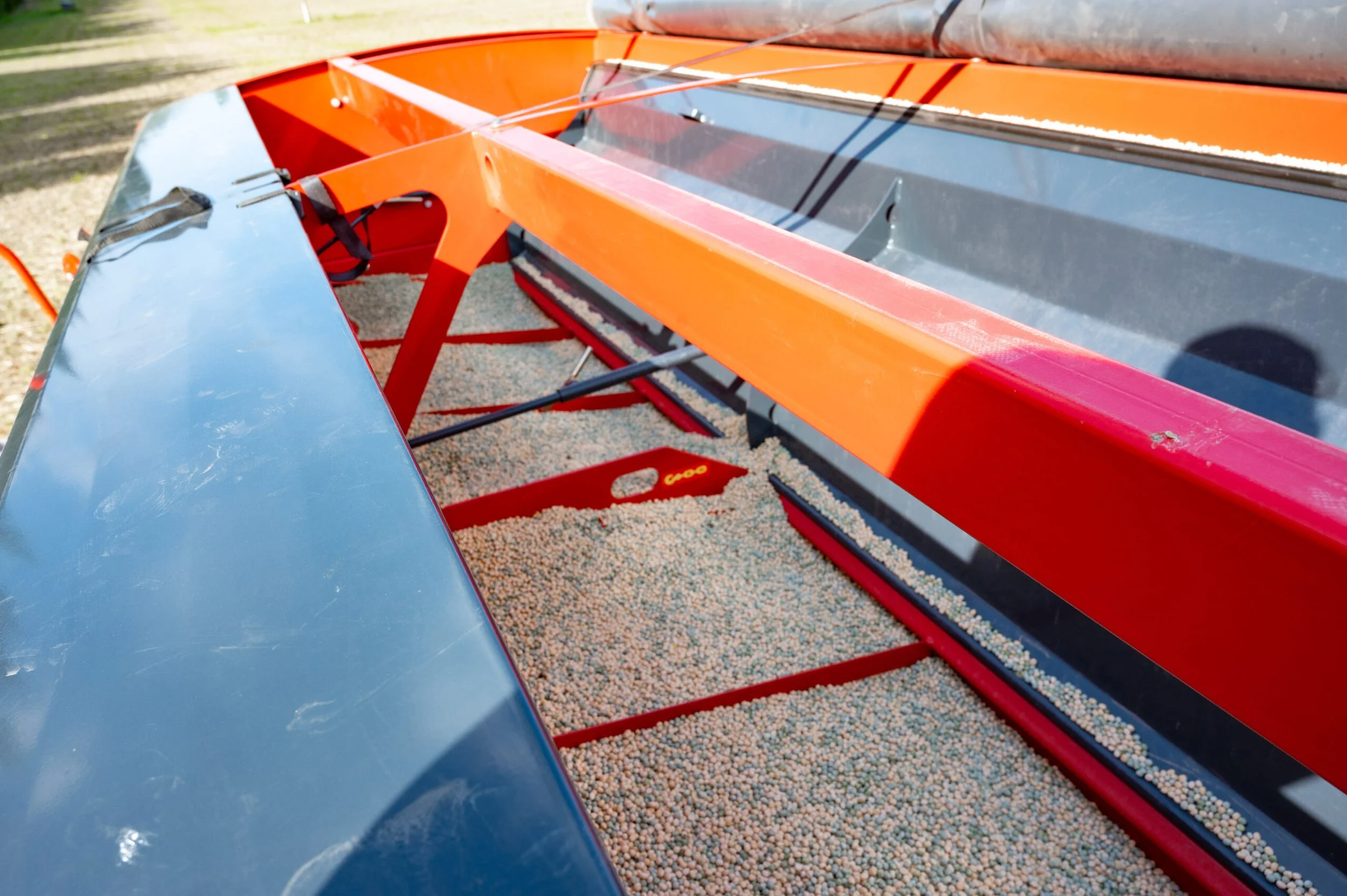 A boat with orange and grey metal parts on a dirt ground, with a blue surface on one side and a large container of small, multicolored pellets or grains underneath.