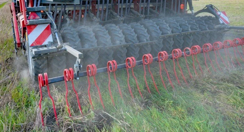 A red and black agricultural machine tilling a field with red tines, kicking up dust and grass.