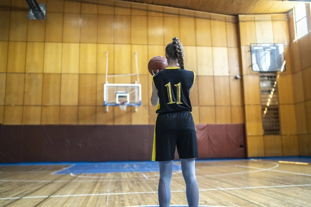A young female basketball player wearing a black jersey with yellow trim and the number 11, holding a basketball in a gymnasium ready to shoot, with a wooden wall and basketball hoop in the background.
