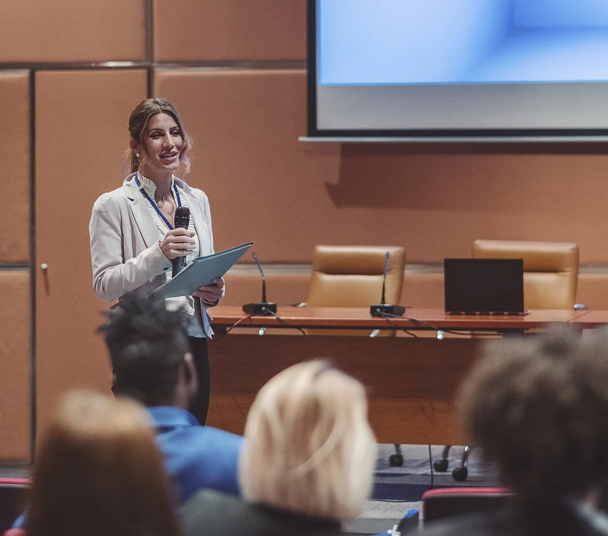 A woman in business attire holding a microphone and notes, standing in front of a conference room with a large screen and chairs, giving a presentation to an audience.