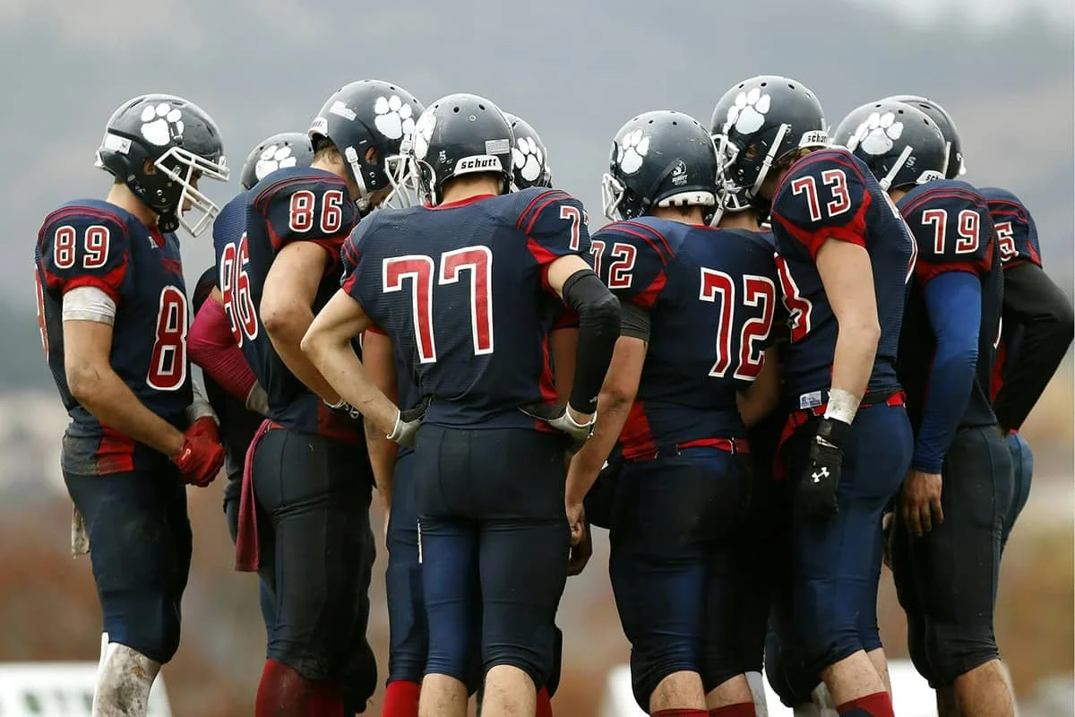A group of football players in navy blue uniforms with red accents, gathered in a huddle on the field, wearing helmets with a white paw print logo, during a game or practice.