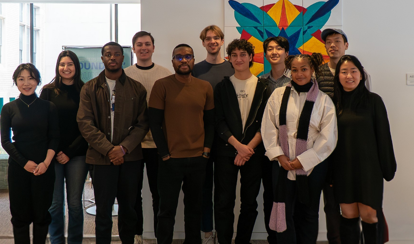 Group of diverse young adults standing together indoors, smiling, with colorful abstract artwork on the wall behind them.