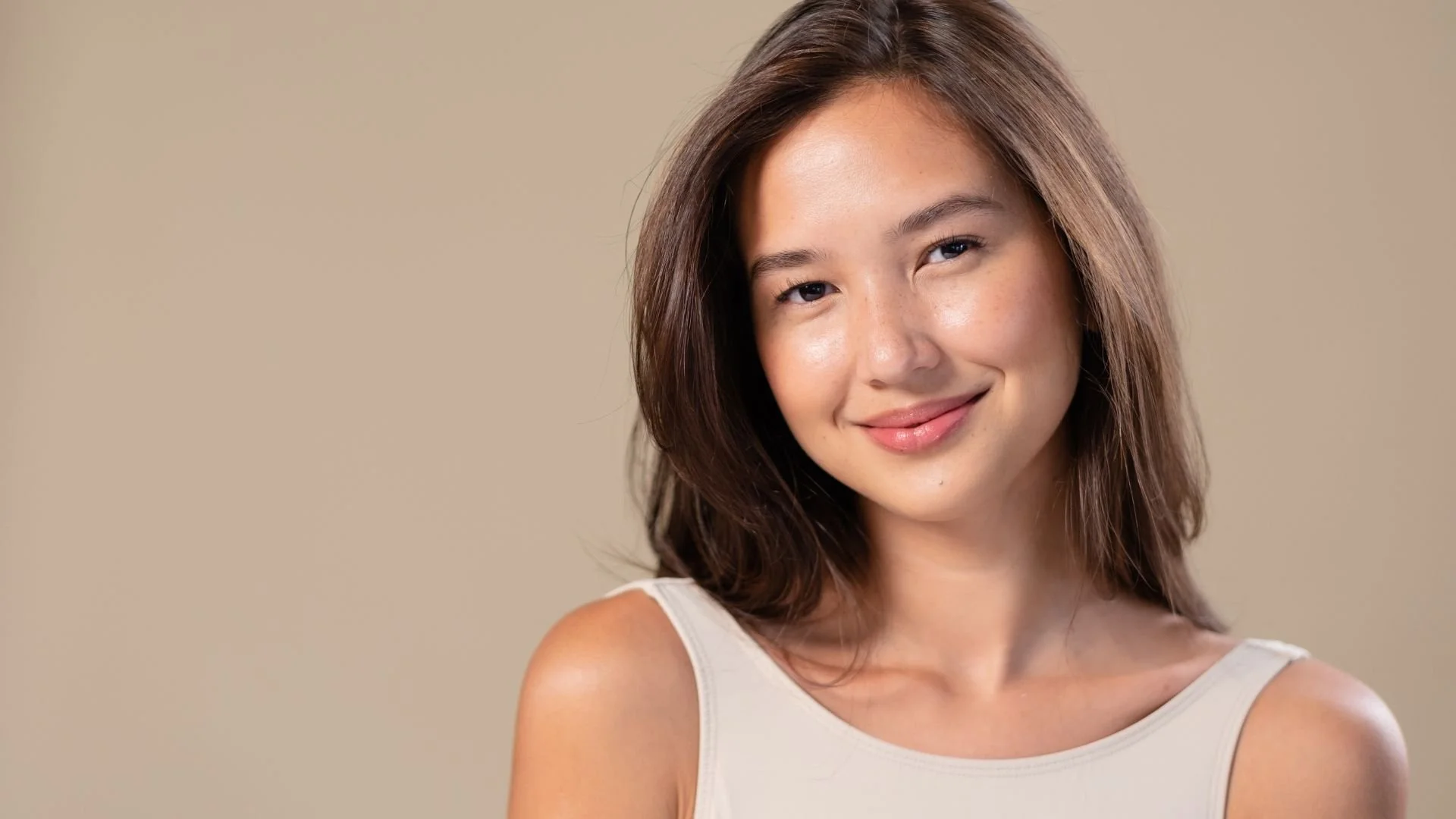 Young woman with shoulder-length brown hair smiling at the camera against a beige background.