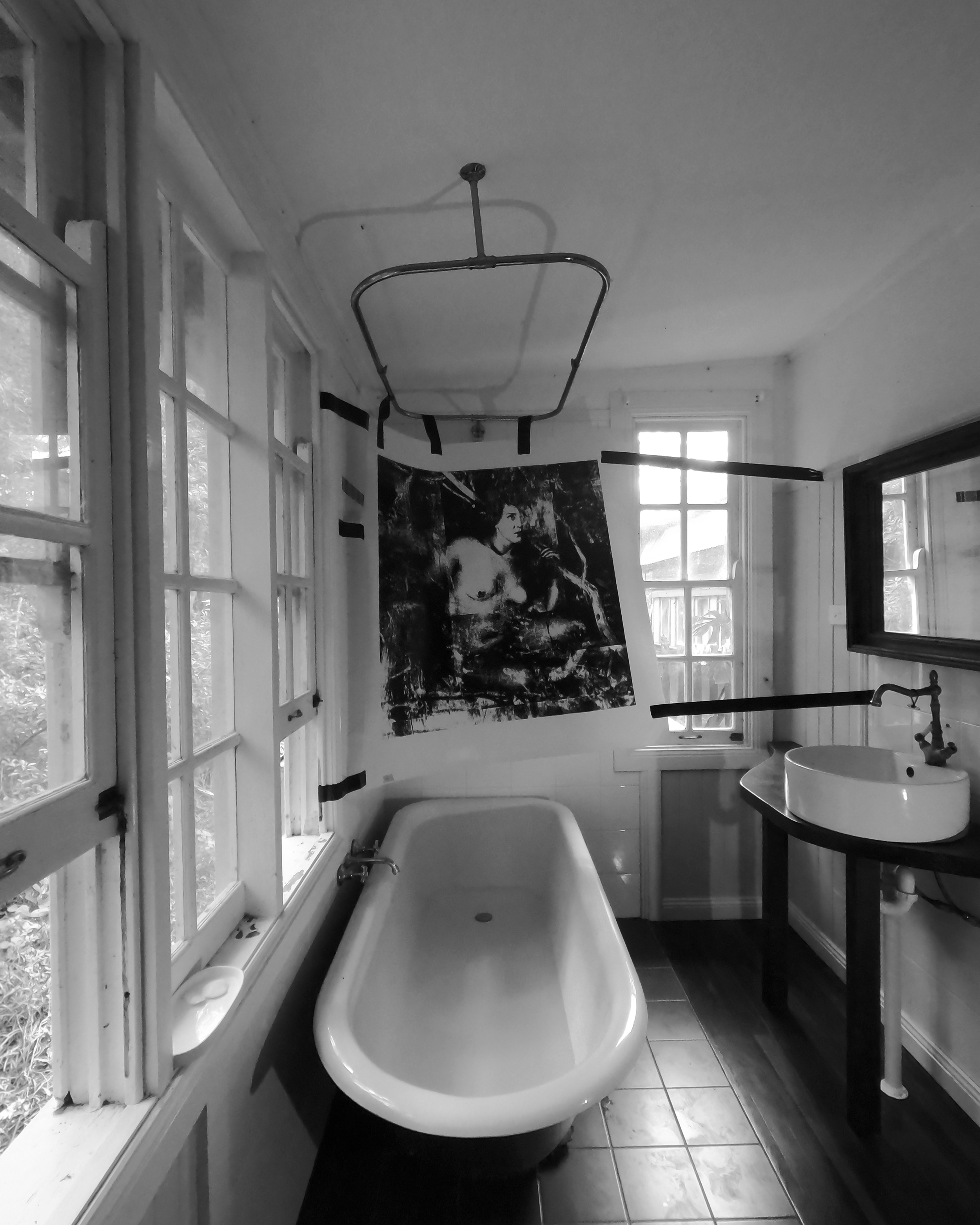 A vintage-style bathroom with a clawfoot bathtub, a sink with a black countertop, large windows with multiple panes, a black photograph of a female figure in a forest-like space is duct taped to the wall, and a ceiling-mounted shower curtain hanger.