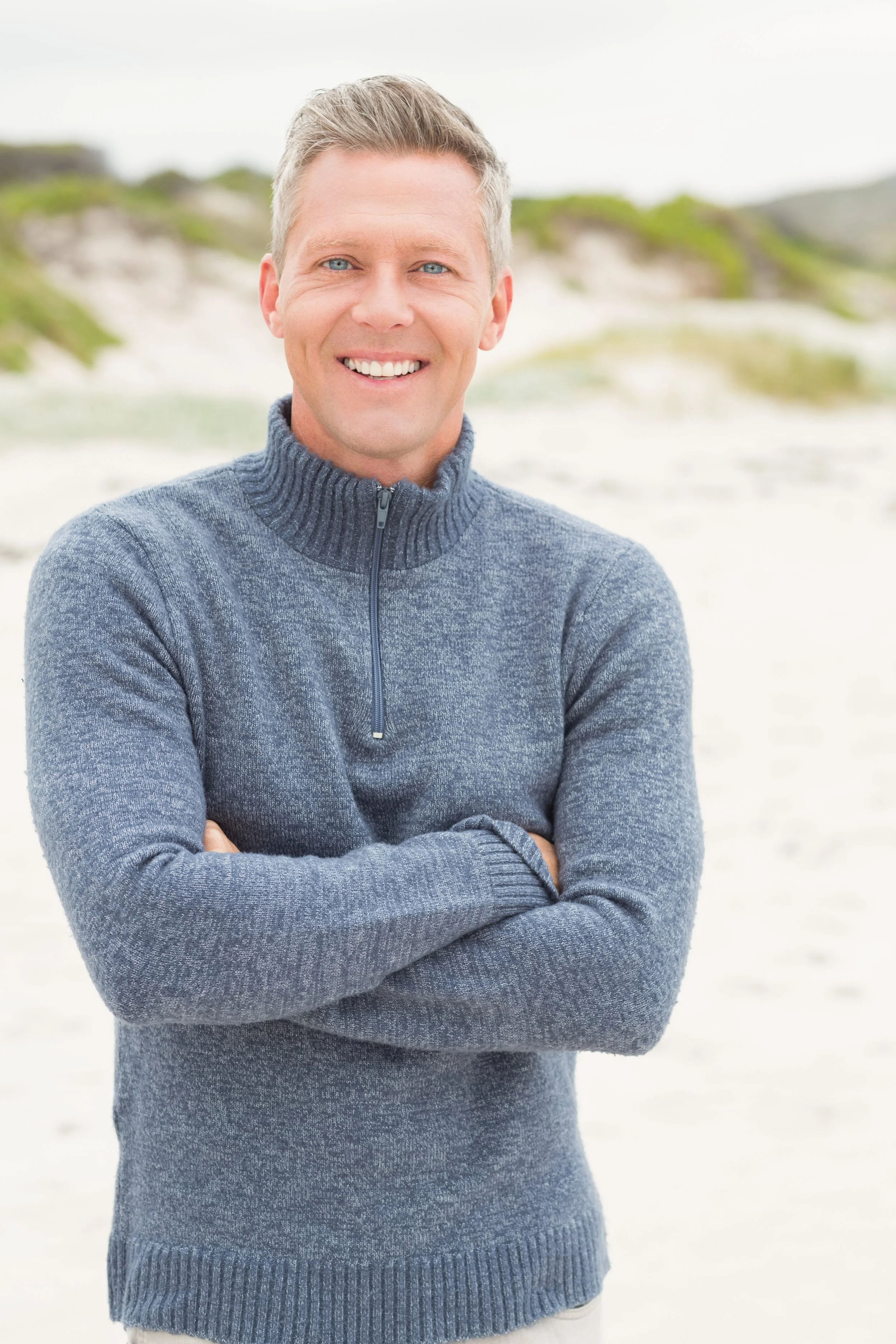 A smiling man with gray hair and blue eyes wearing a gray zip-up sweater, standing on a sandy beach with dunes in the background.