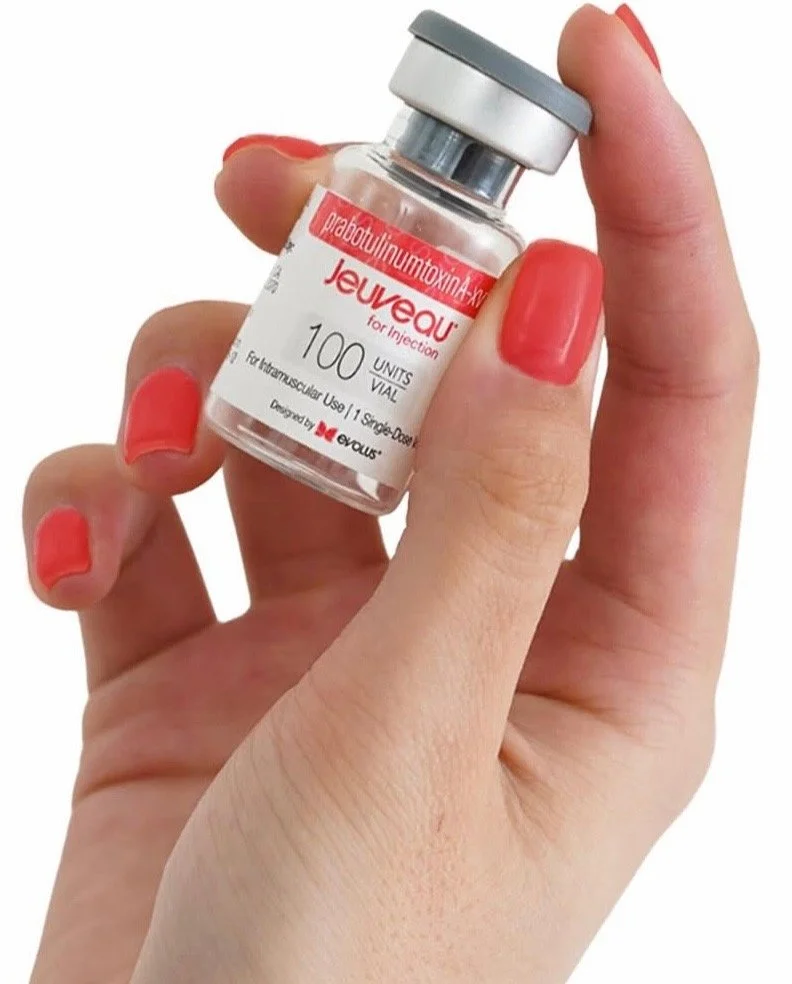 A woman with closed eyes receiving a facial treatment, with a skincare brush applying product to her face, on a treatment bed with a white headband.