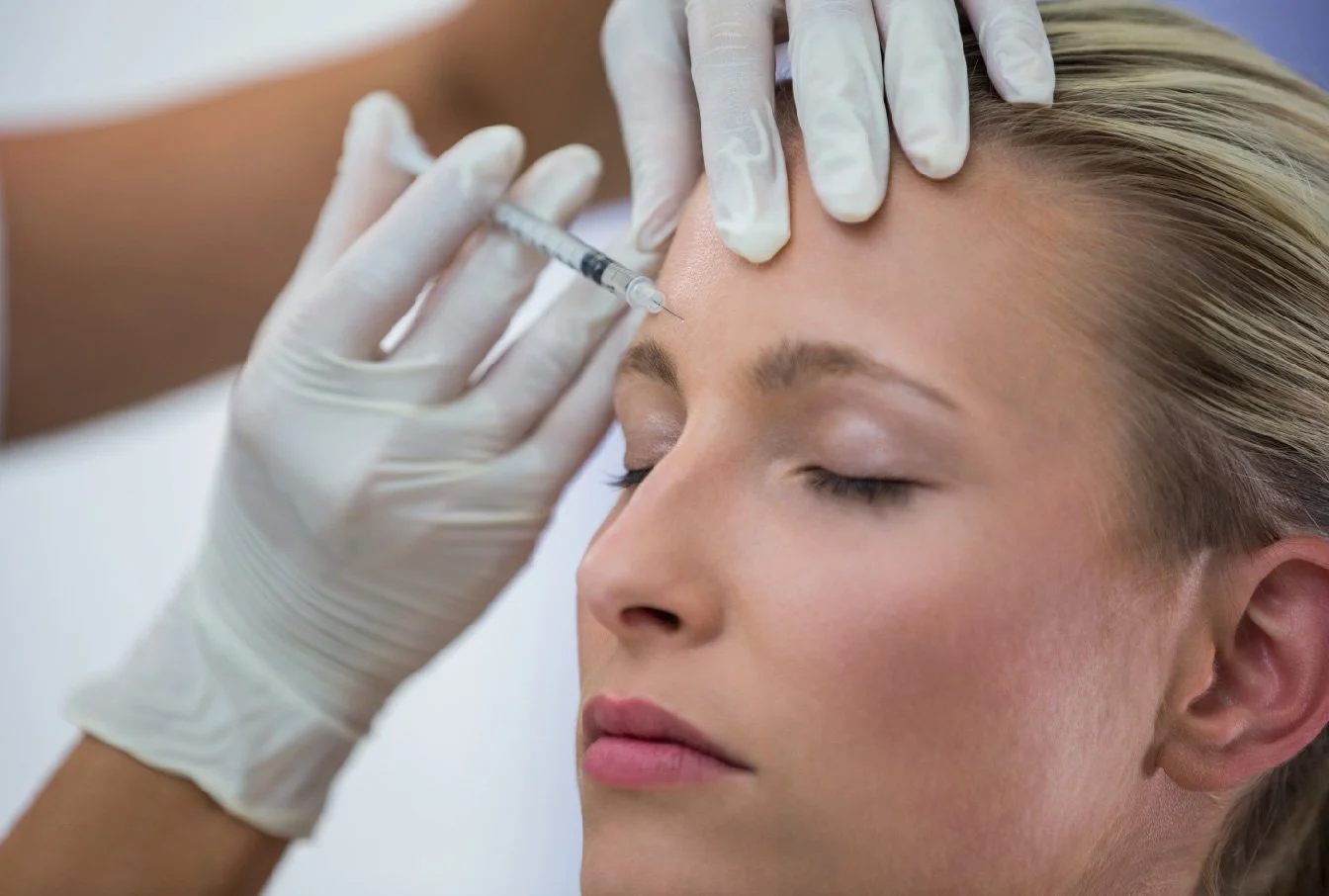A woman with closed eyes receives an injection in her forehead from a medical professional wearing gloves.