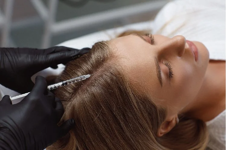 A woman receiving a cosmetic injection in her scalp from a medical professional wearing black gloves.