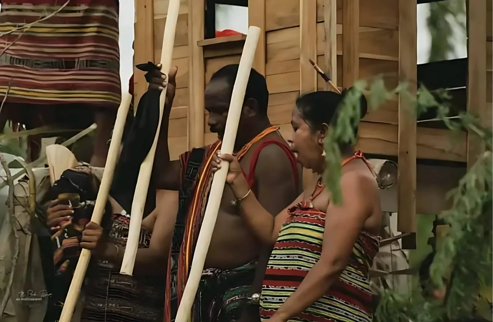 Two indigenous individuals wearing traditional striped clothing, holding wooden sticks, standing in front of a wooden structure, engaging in a cultural activity.