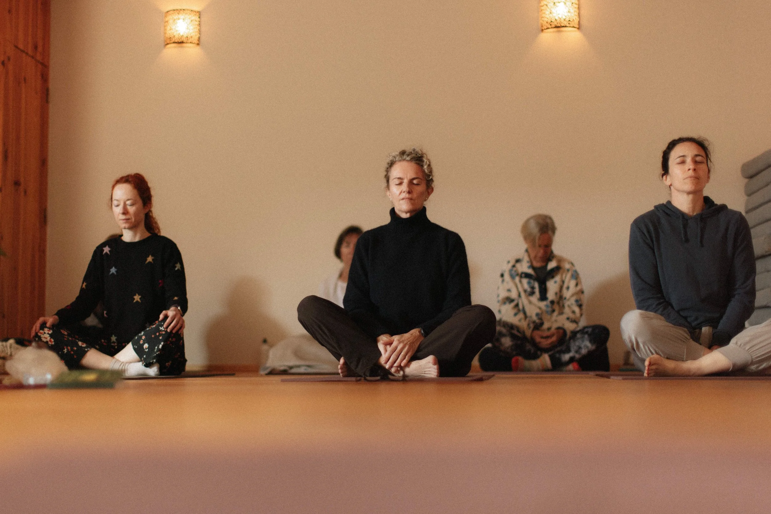 Group of five women sitting cross-legged on a wooden deck outdoors, under a large tree, with a background of bushes and clear blue sky, participating in a mindfulness session.