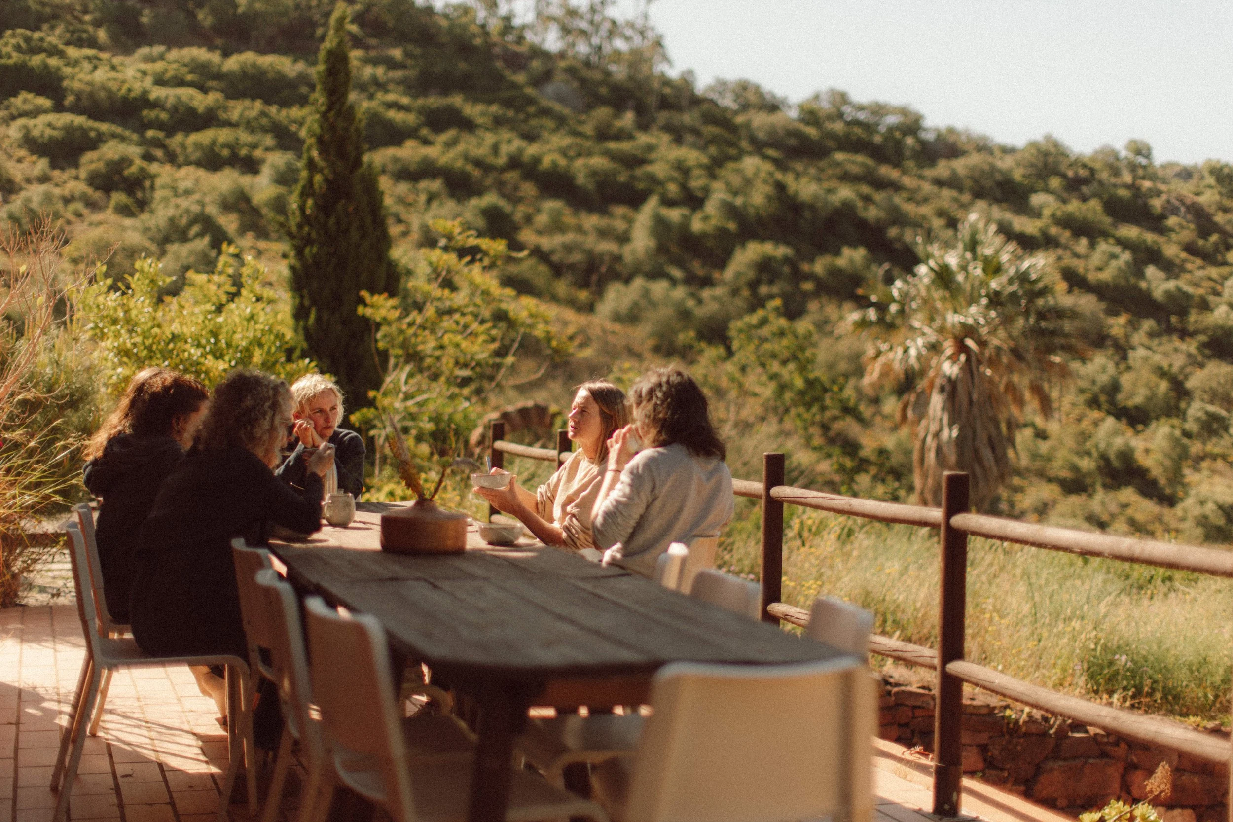 A group of people sitting around a wooden picnic table outdoors during sunset, with a large beige shade sail overhead and rolling hills in the background.