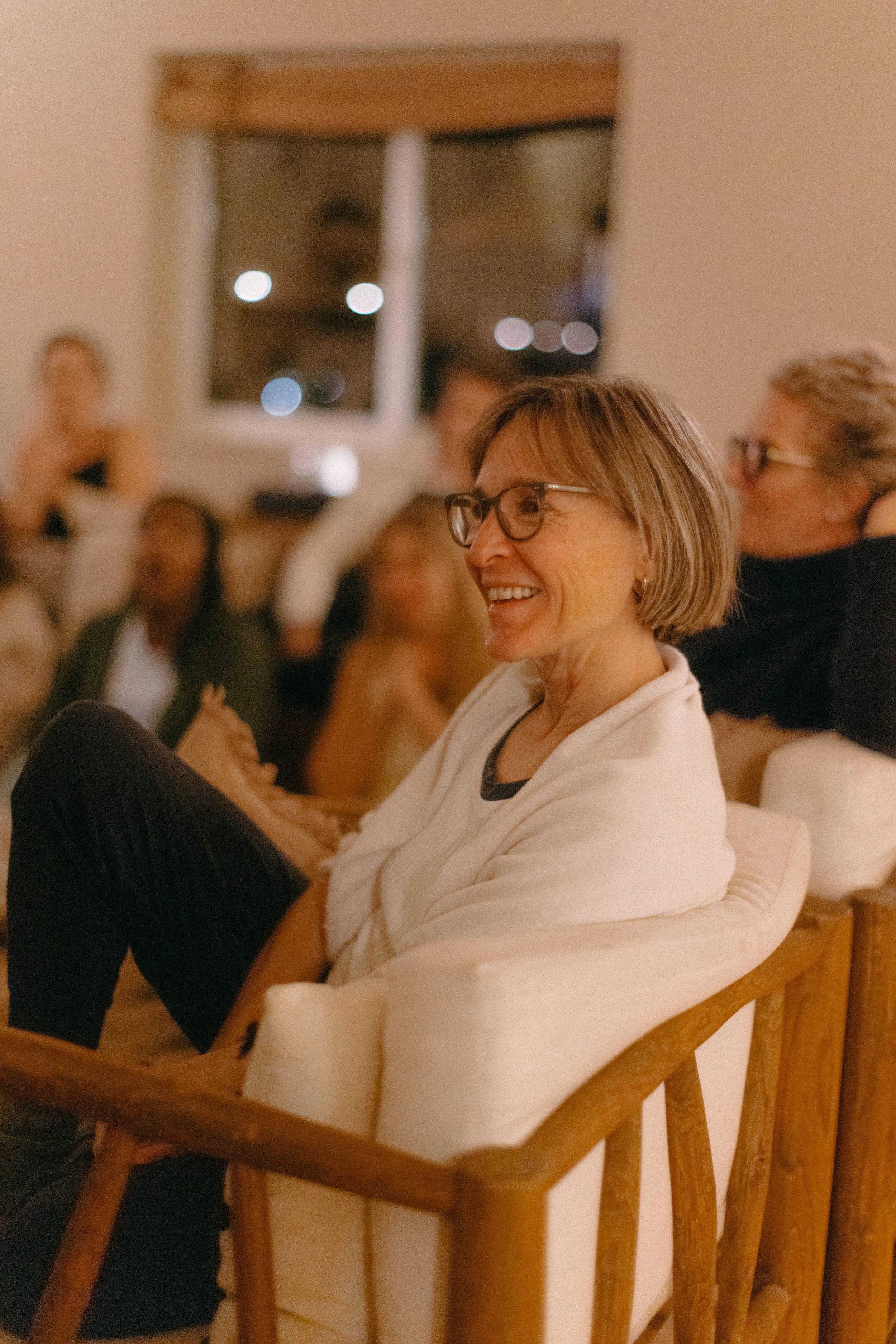 Two women sitting on a cozy rug, one singing with hand on chest and the other playing guitar, in a warmly lit room decorated with spiritual items on a side table.