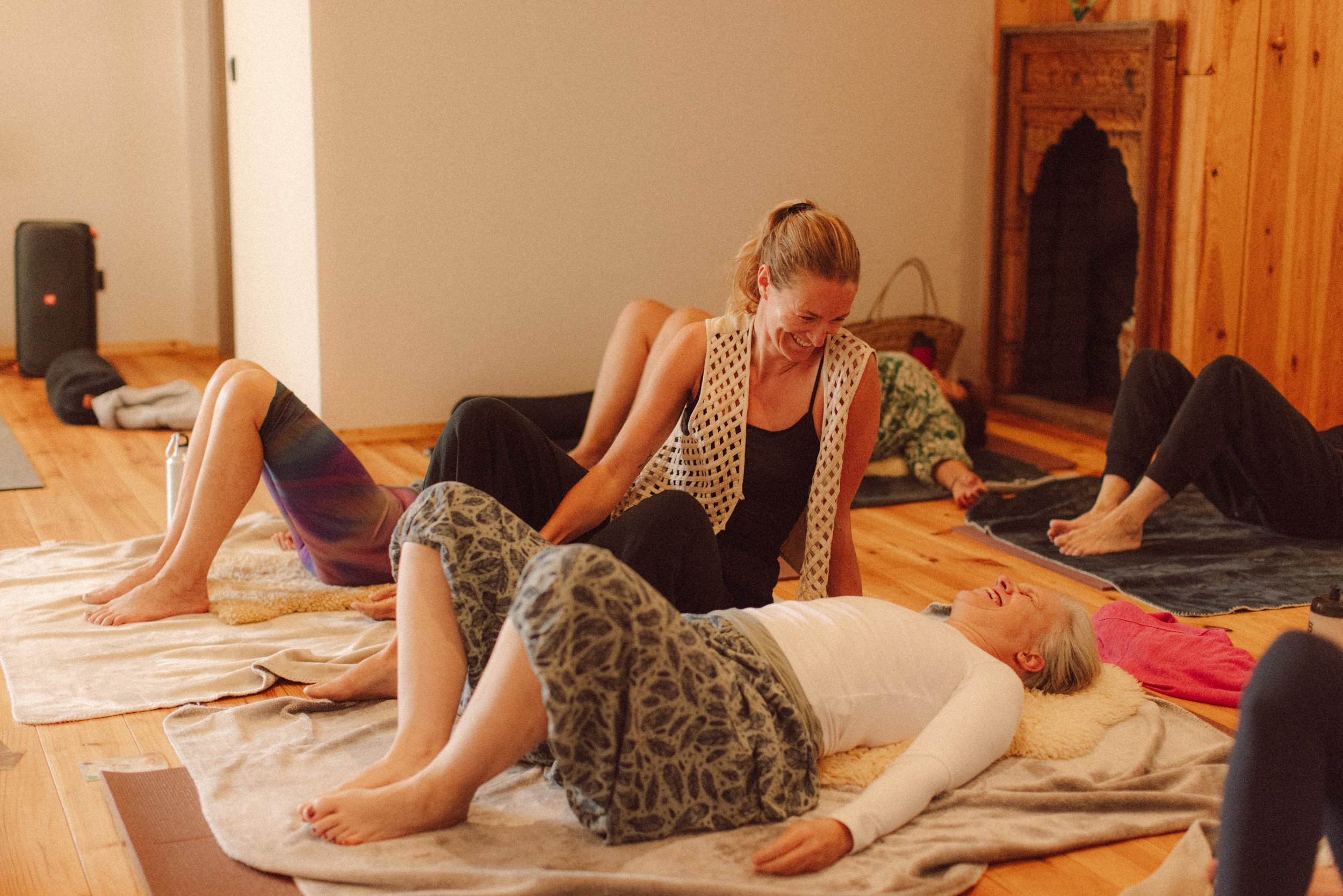 A group of women sitting cross-legged on the floor inside a wooden pavilion with large windows, participating in a meditation or spiritual gathering.