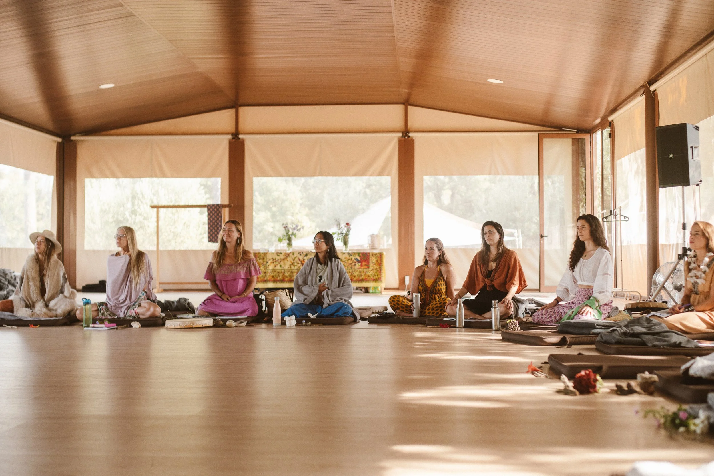 A group of women sitting cross-legged on the floor inside a wooden pavilion with large windows, participating in a meditation or spiritual gathering.