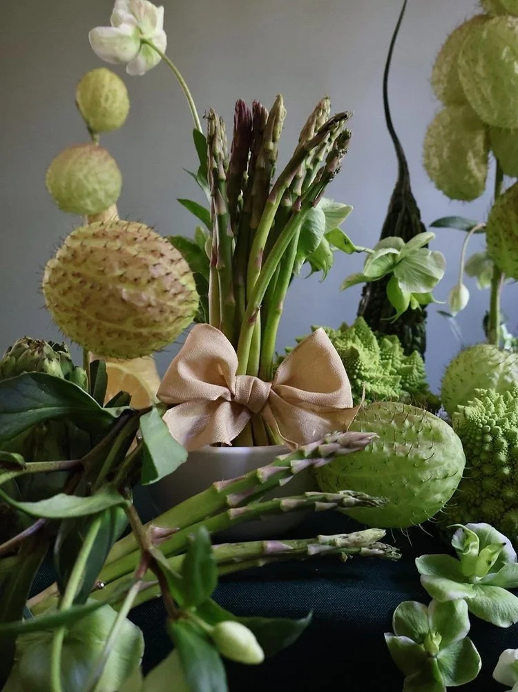 A potted arrangement of various green plants and flowers, including asparagus, pincushion protea, and succulents, with a beige bow around the asparagus stalks.