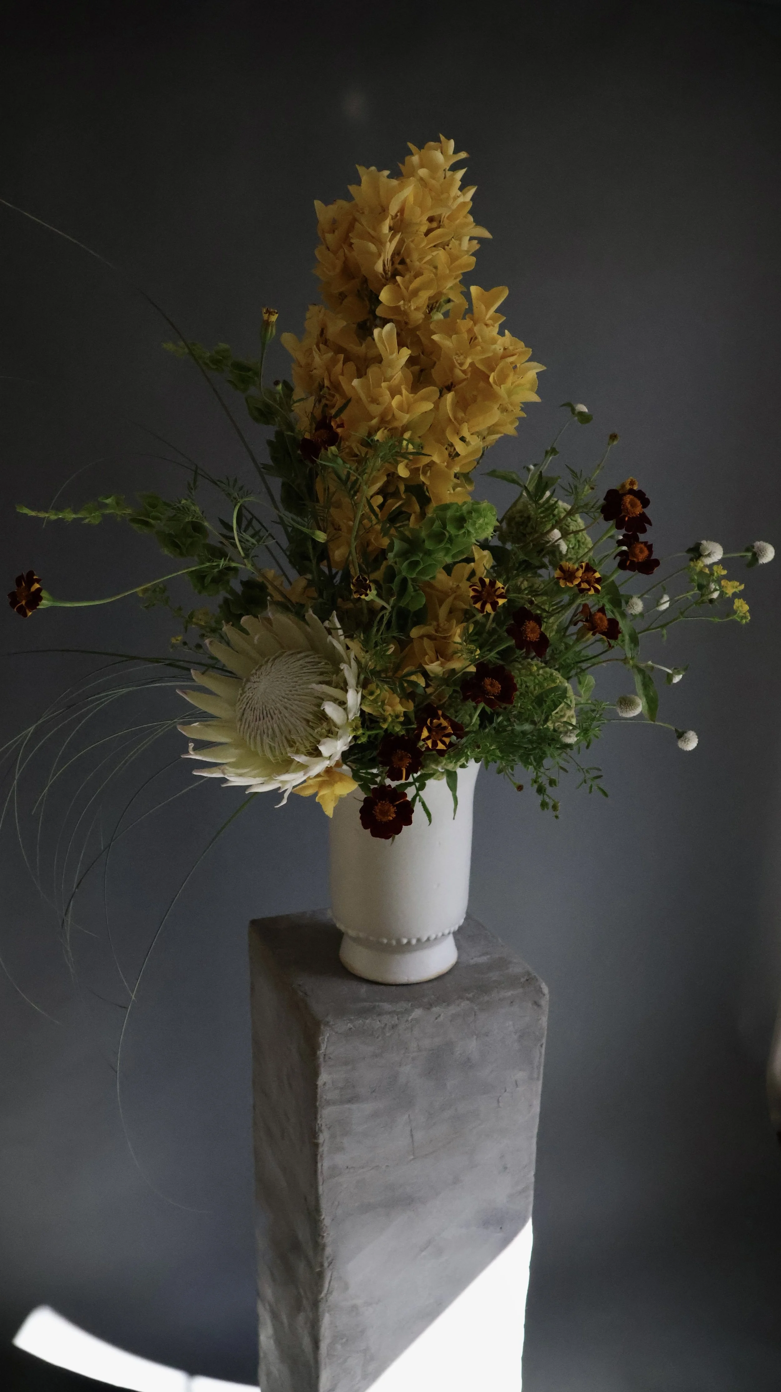 A tall beige vase with a floral arrangement of yellow, white, and dark red flowers on a concrete pedestal against a dark background.