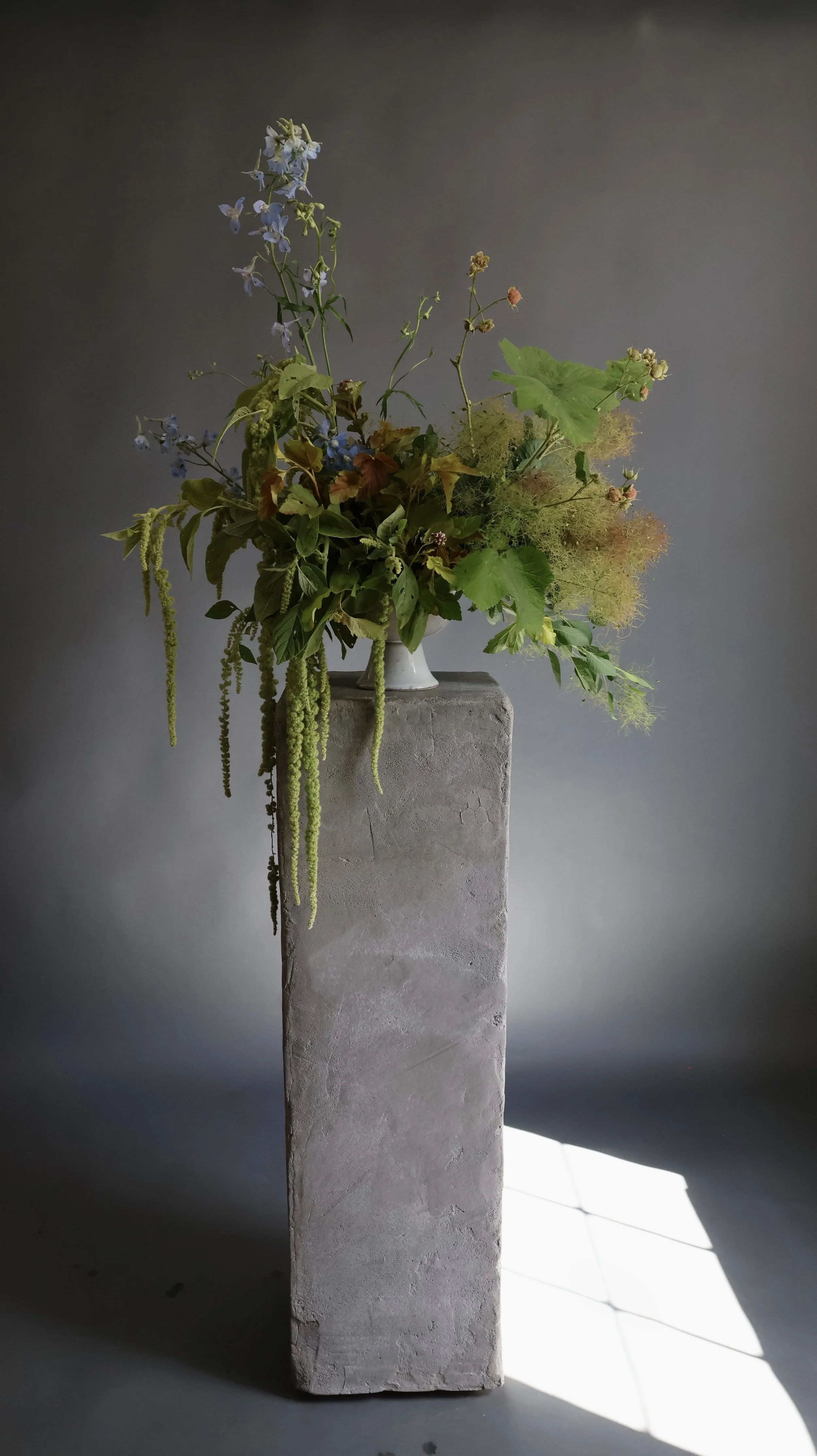 A flower arrangement in a white vase atop a tall concrete pedestal, with various green leaves and hanging green flowers against a dark background, illuminated by sunlight.