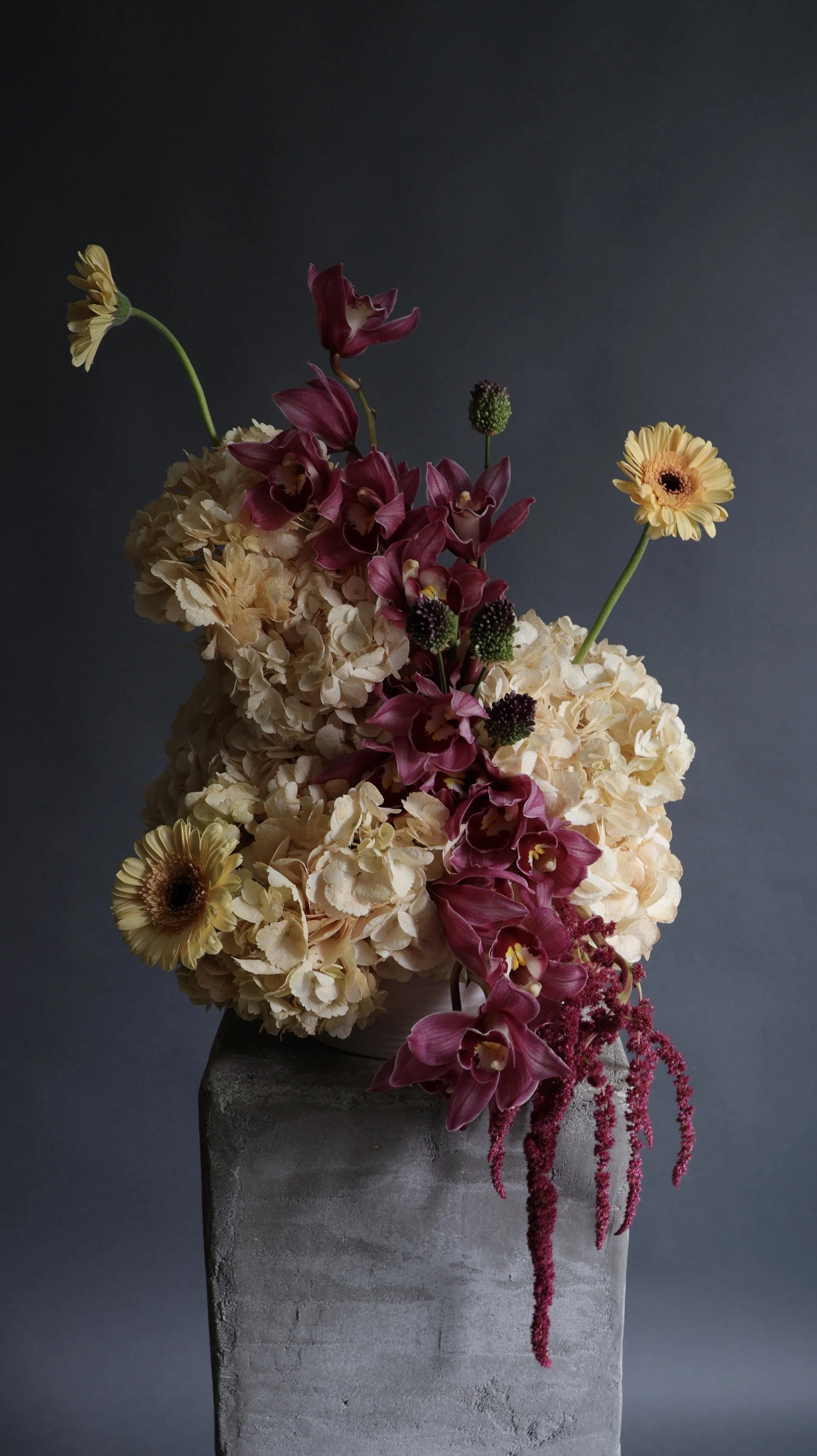 A floral arrangement with cream hydrangeas, deep pink orchids, yellow gerbera daisies, and pink astilbe in a gray vase against a dark background.