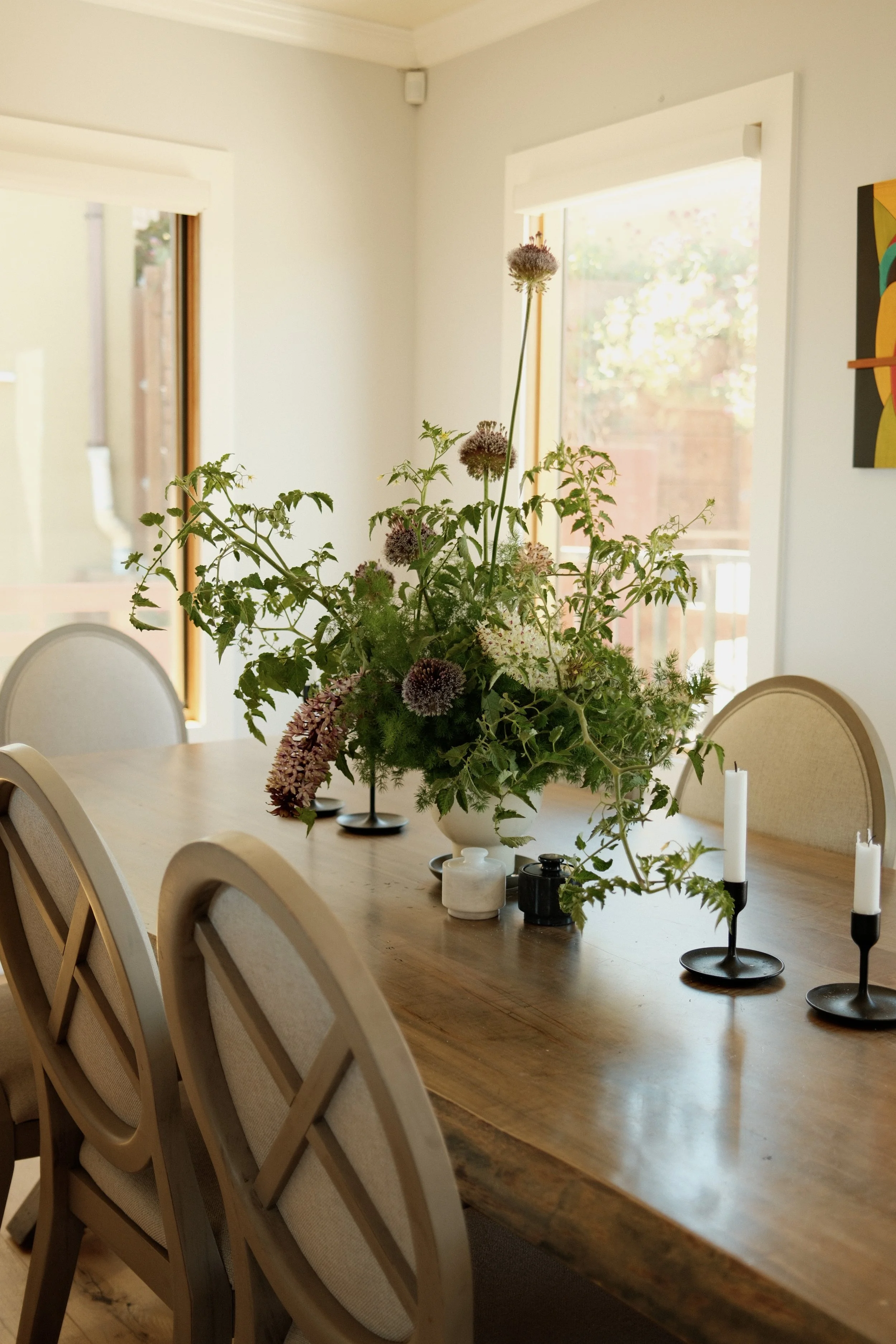 A dining room table with a large floral arrangement, black and white candles, and small decorative vases, with windows in the background.