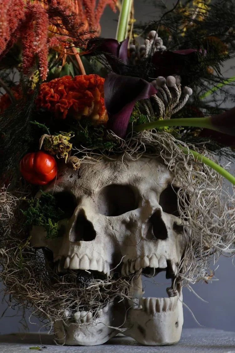 A skull styled with dried flowers and plants, including a red flower, small mushrooms, and next to a red tomato.